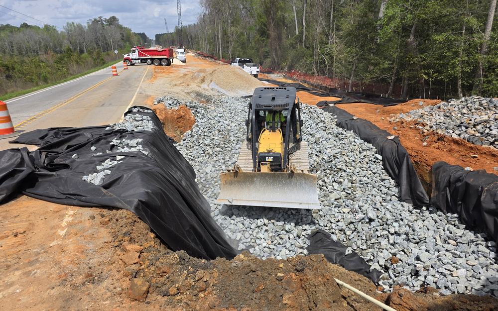 A compact excavator placing stone on a highway shoulder beside erosion-control matting