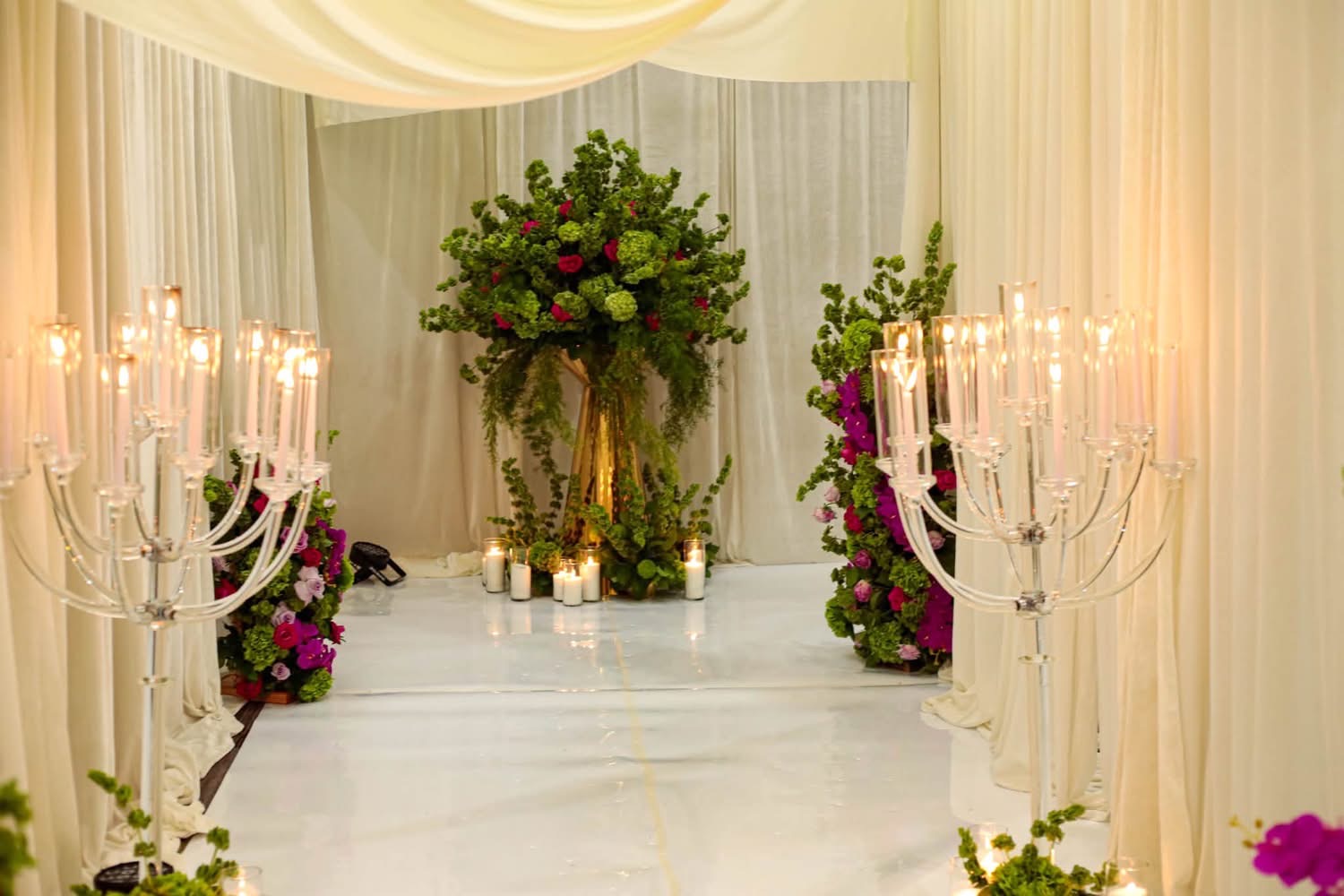 Opulent white gloss aisle lined with ivory drapery, multi-tiered crystal candelabras with taper candles, and tall floral centerpieces