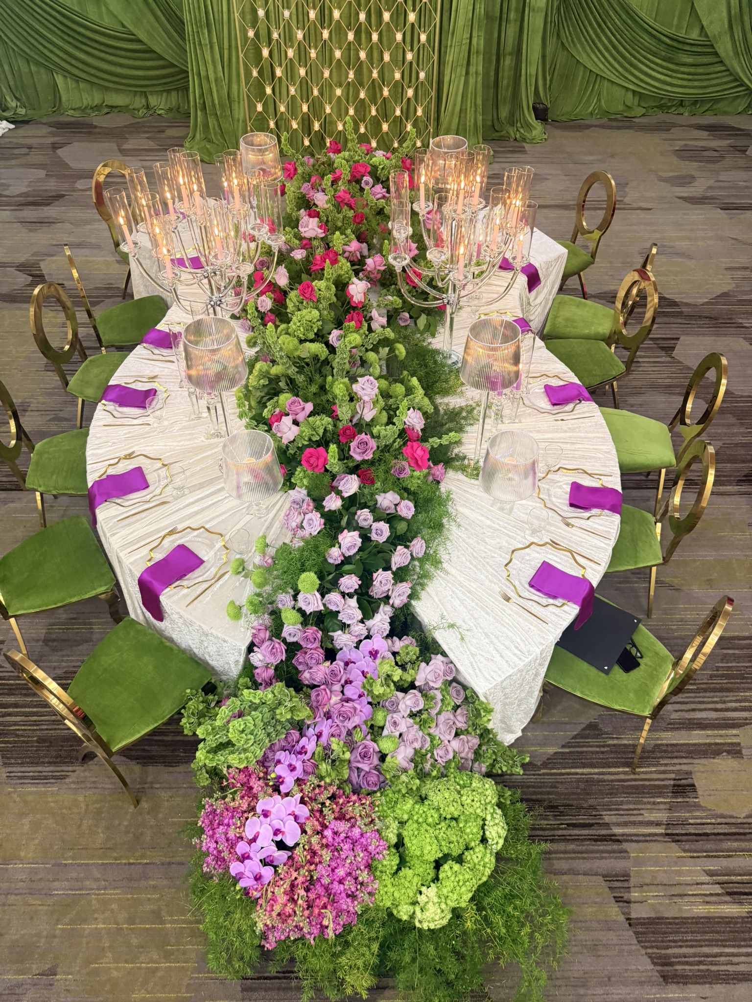 Luxurious oval banquet table draped in satin with cascading purple, blush, and green florals, velvet green chairs, and towering crystal candelabras