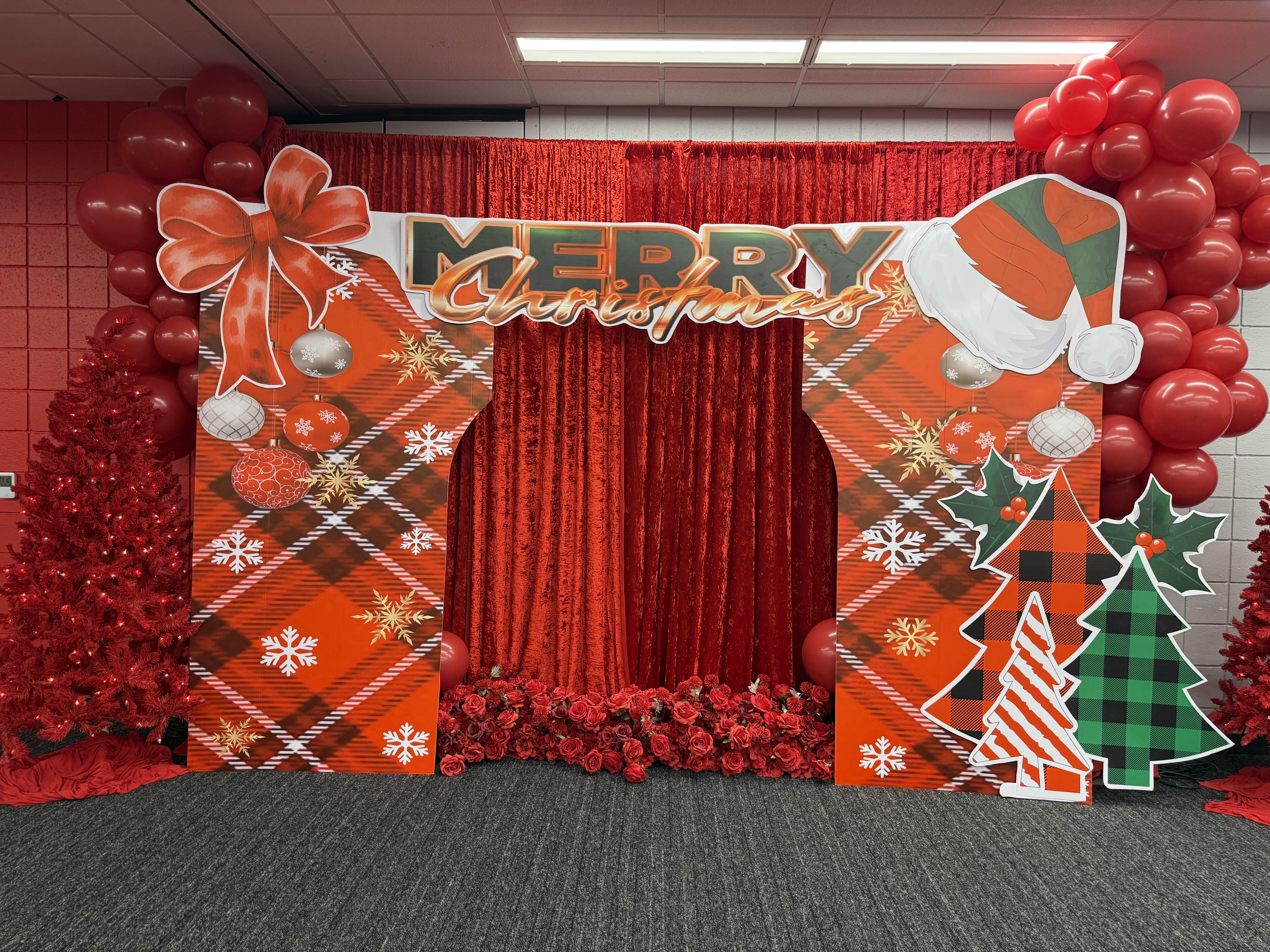 Holiday photo arch with festive red plaid framing, 'Merry Christmas' header, rich red velvet backdrop, red balloon clusters, and lit red Christmas trees