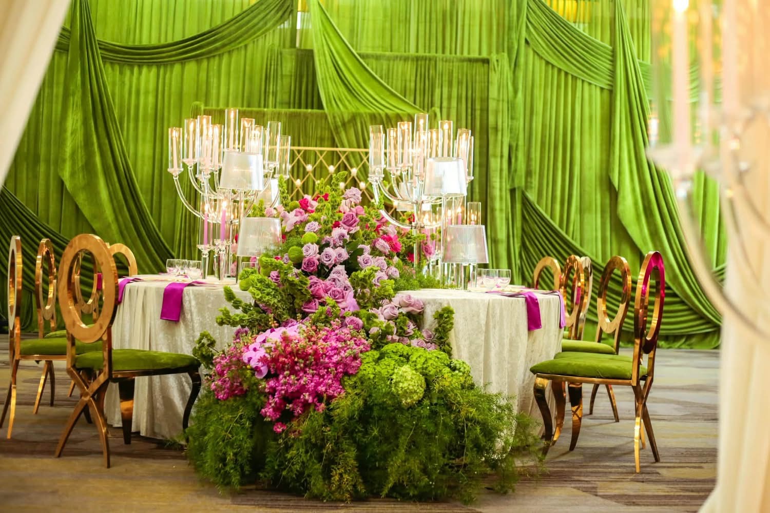 Grand luxury banquet hall with draped lime velvet curtains, overflowing purple and green floral table runner, and glowing crystal candelabras