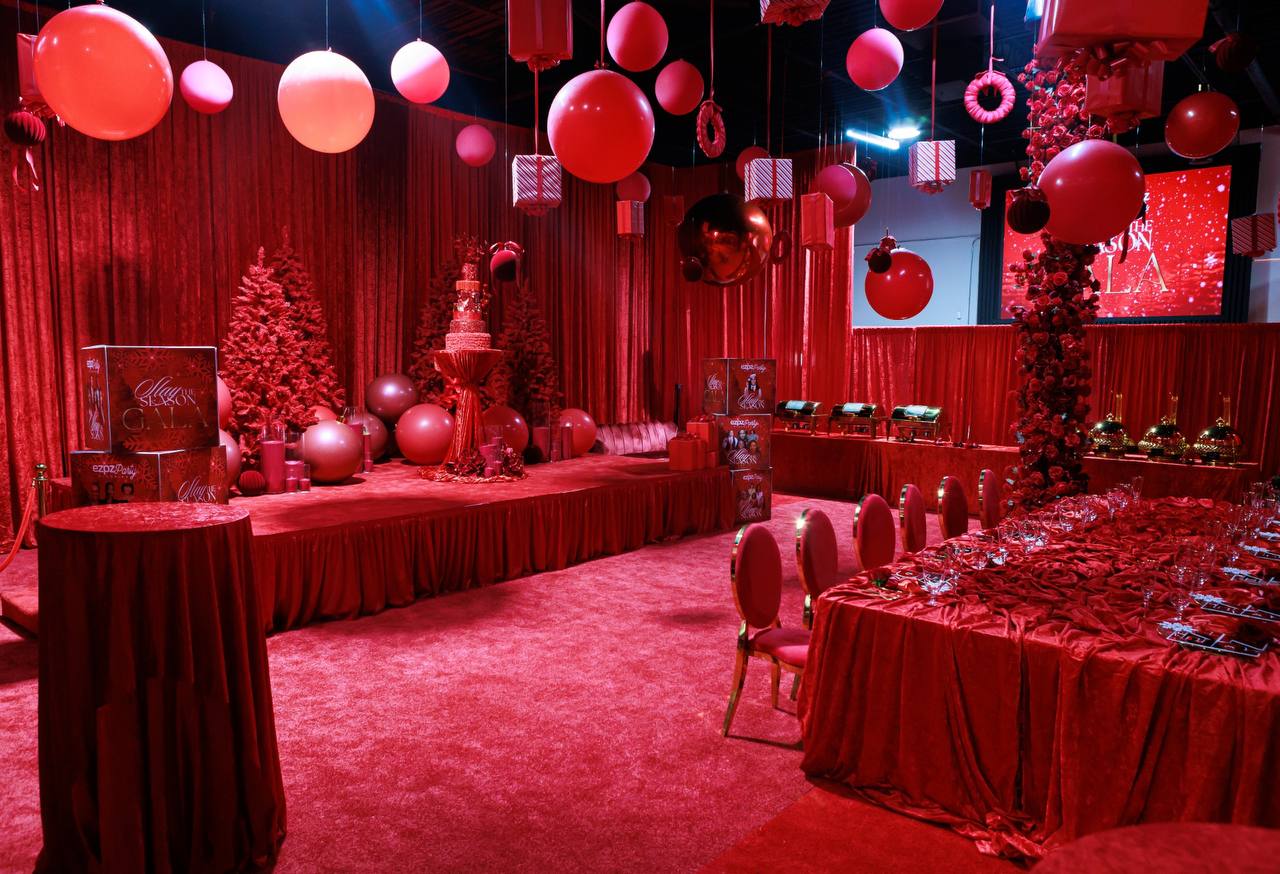 Full room view of an opulent all-red holiday gala featuring velvet draping, crimson stage, red Christmas trees, floating balloon spheres, and tablescapes