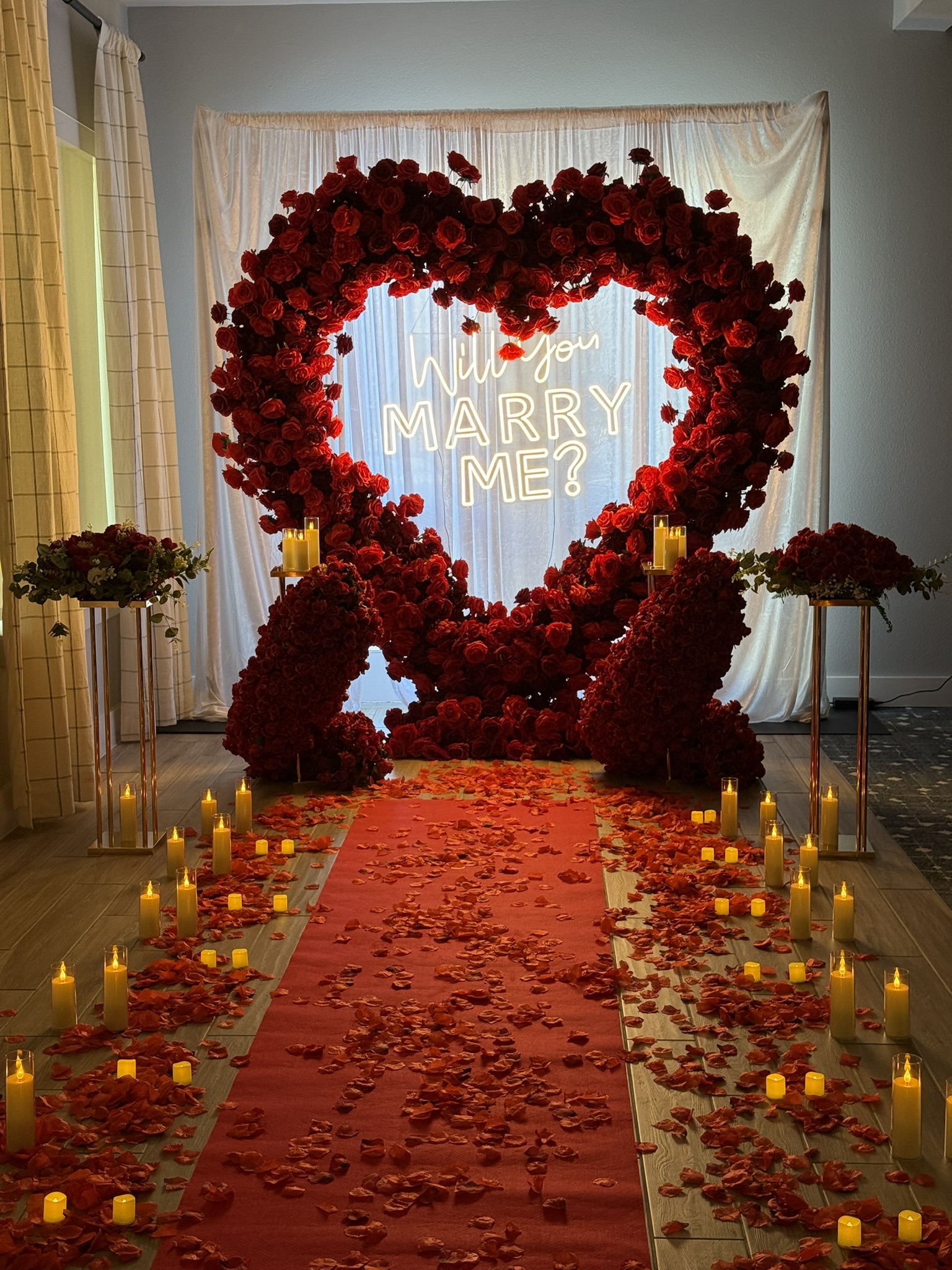 Romantic red rose floral heart installation with 'Will You Marry Me?' neon sign and candlelit aisle