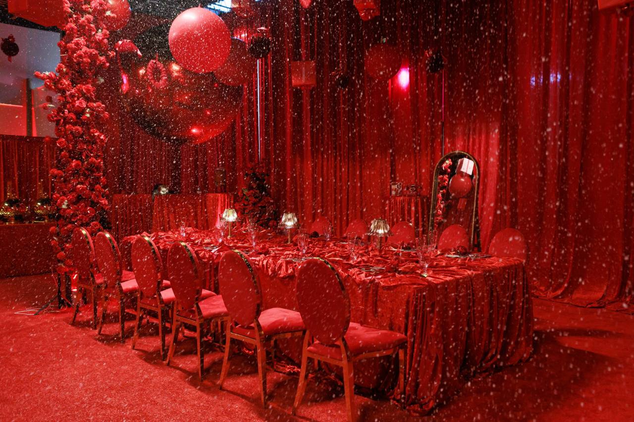 Luxe banquet table draped in crushed red velvet with crystal lamps, gold chairs, and simulated falling snow under red drapery