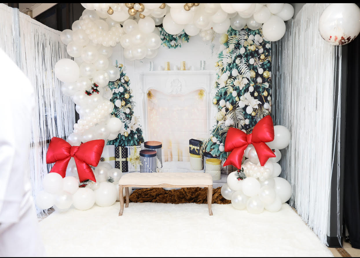 Winter wonderland holiday setup featuring a lush white and gold balloon garland, satin red bows, cozy lit fireplace backdrop, and bench