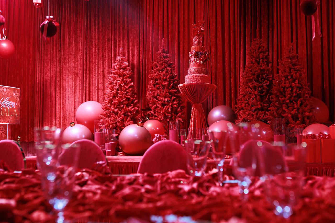 Close-up of crimson velvet gala stage styling with centerpiece tiered cake, crimson Christmas trees, and oversized metallic ornaments