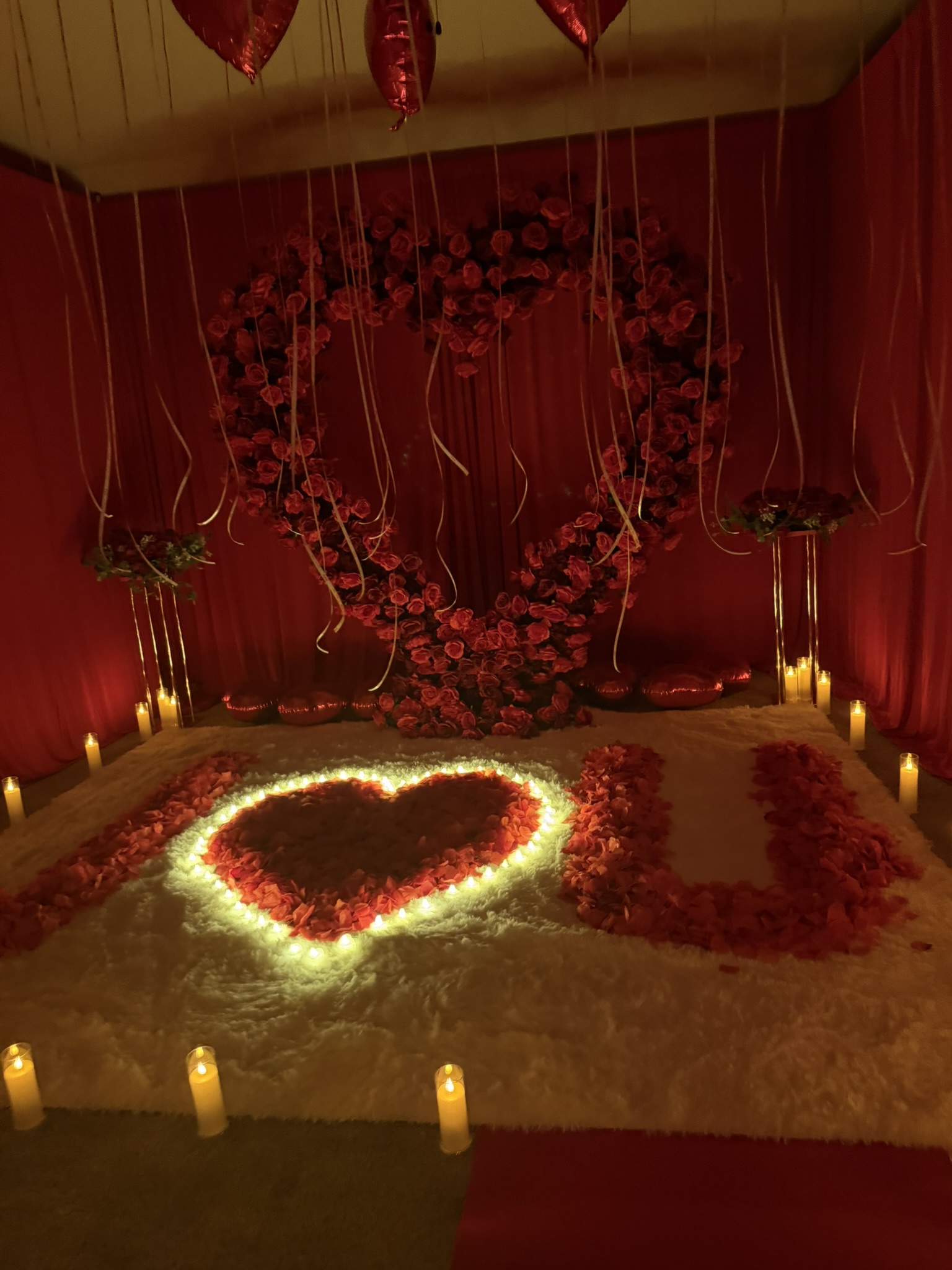 Intimate proposal styling featuring giant red rose heart arch, illuminated candlelit rose petal 'I Heart U' display, and plush white rug