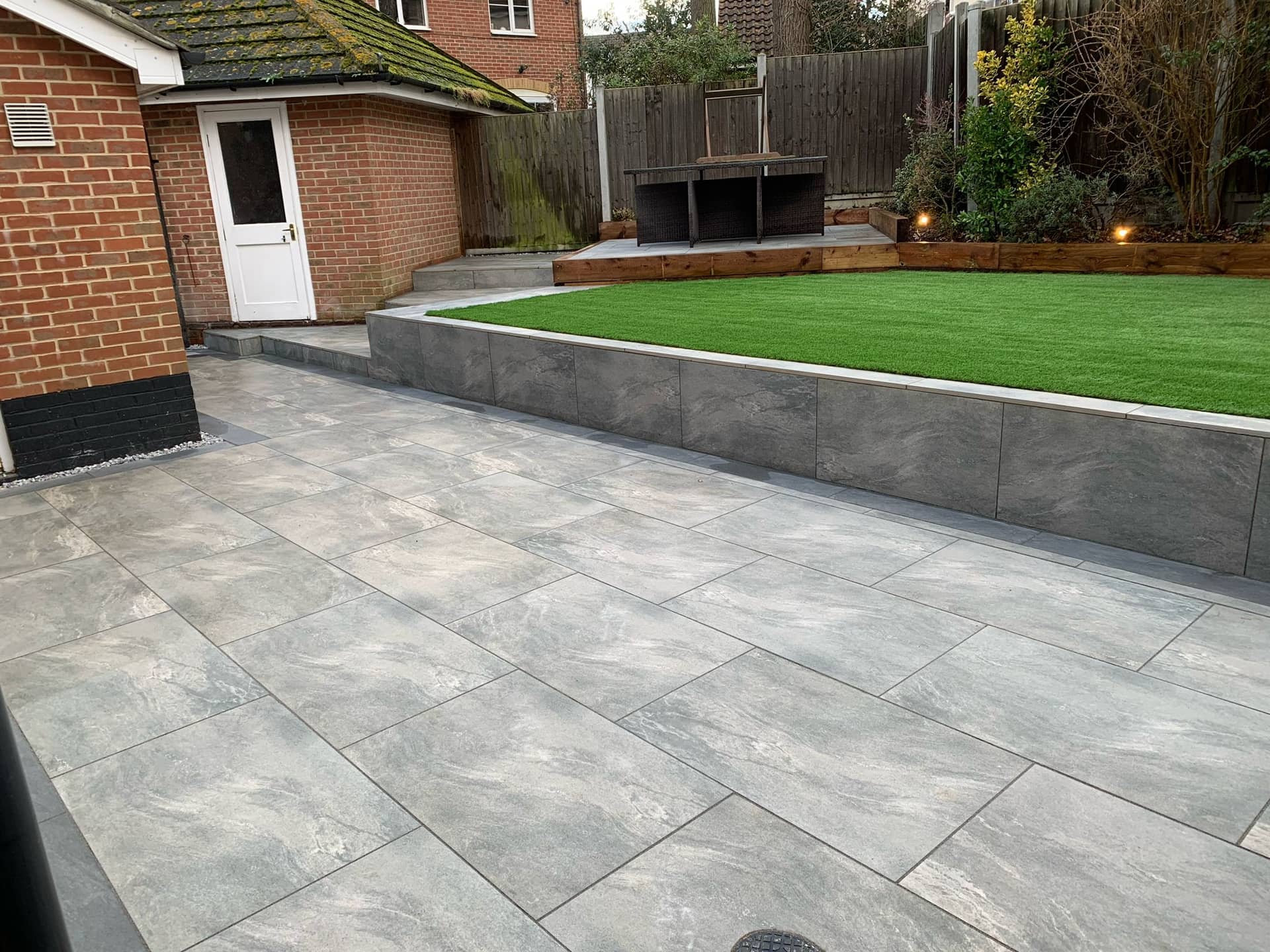 Raised artificial grass lawn tier with grey porcelain patio paving and sleeper-edged planter beds