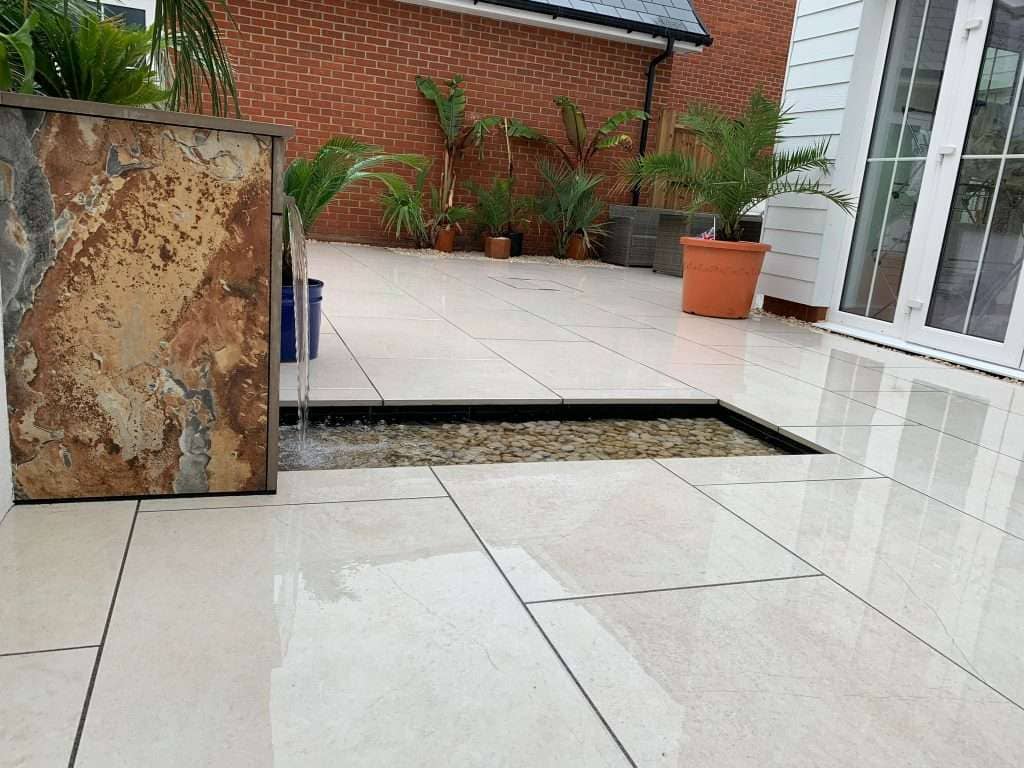 Stone water feature spilling into a pebble-lined drainage channel on cream porcelain paving in Rayleigh