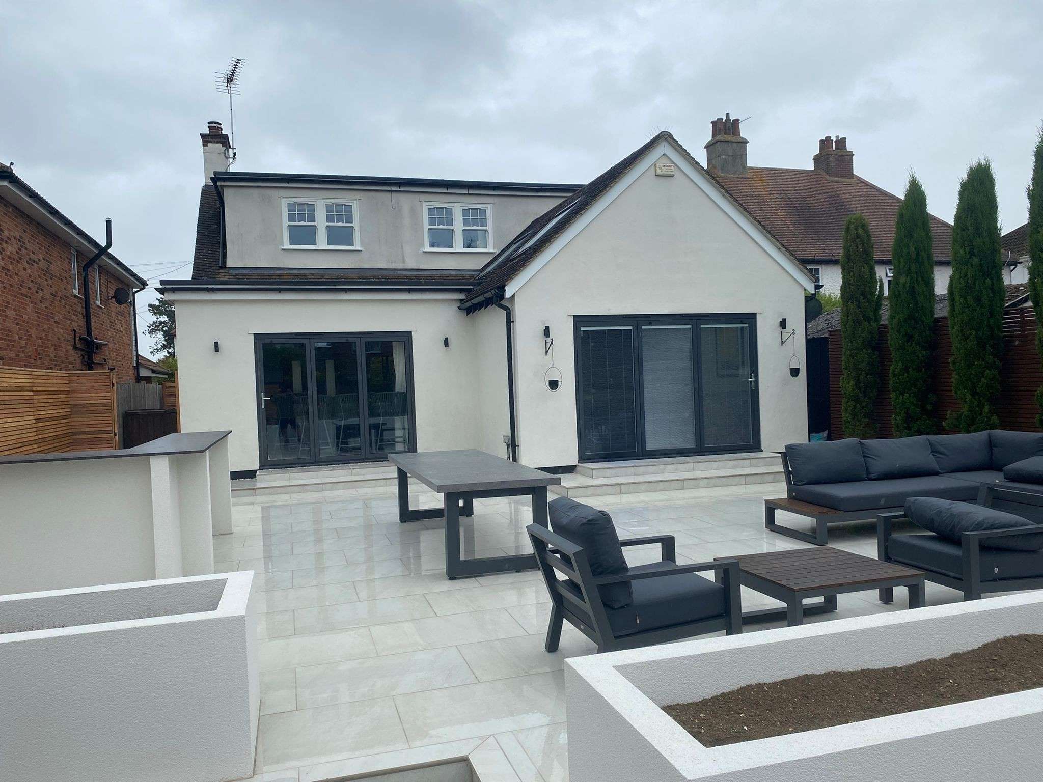 Whole garden transformation in Hadleigh showing light porcelain patio with outdoor dining table, lounge seating, outdoor bar structure and rendered planter beds