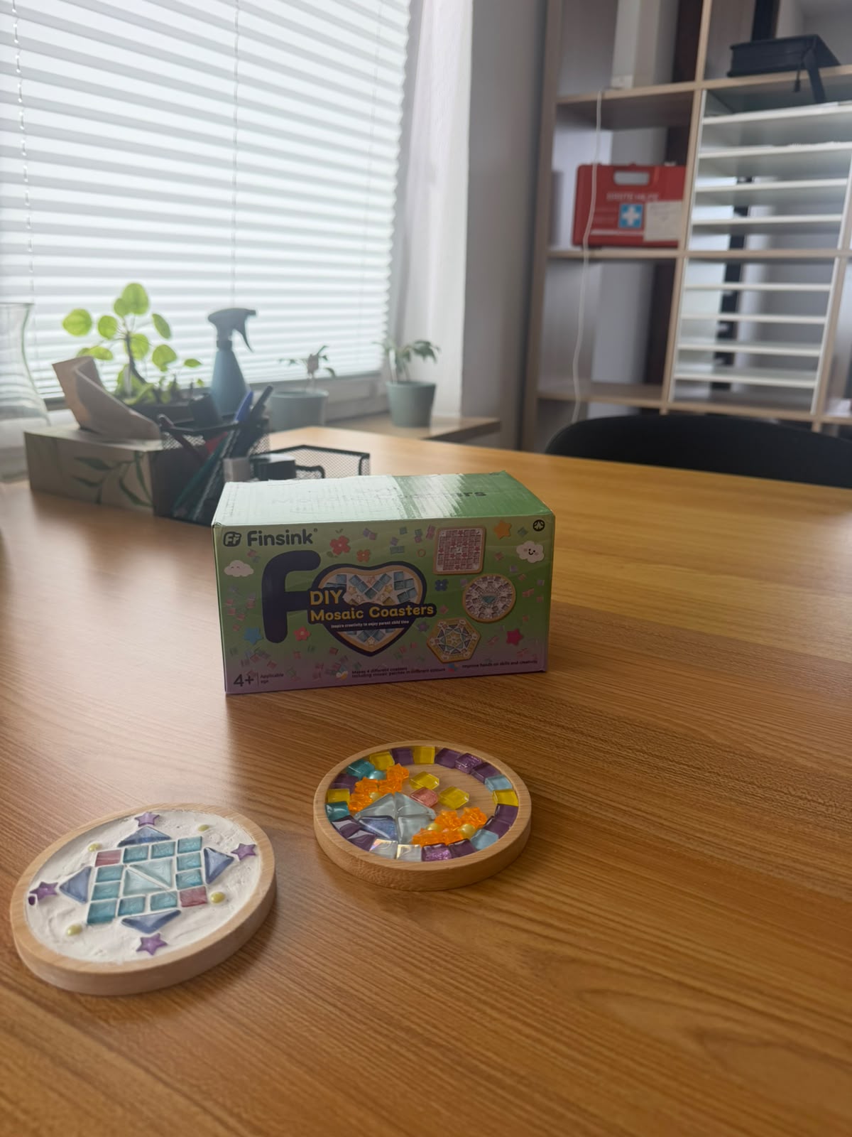 Zwei fertige Mosaik-Untersetzer aus Glassteinen vor einer Bastelschachtel auf einem Holztisch