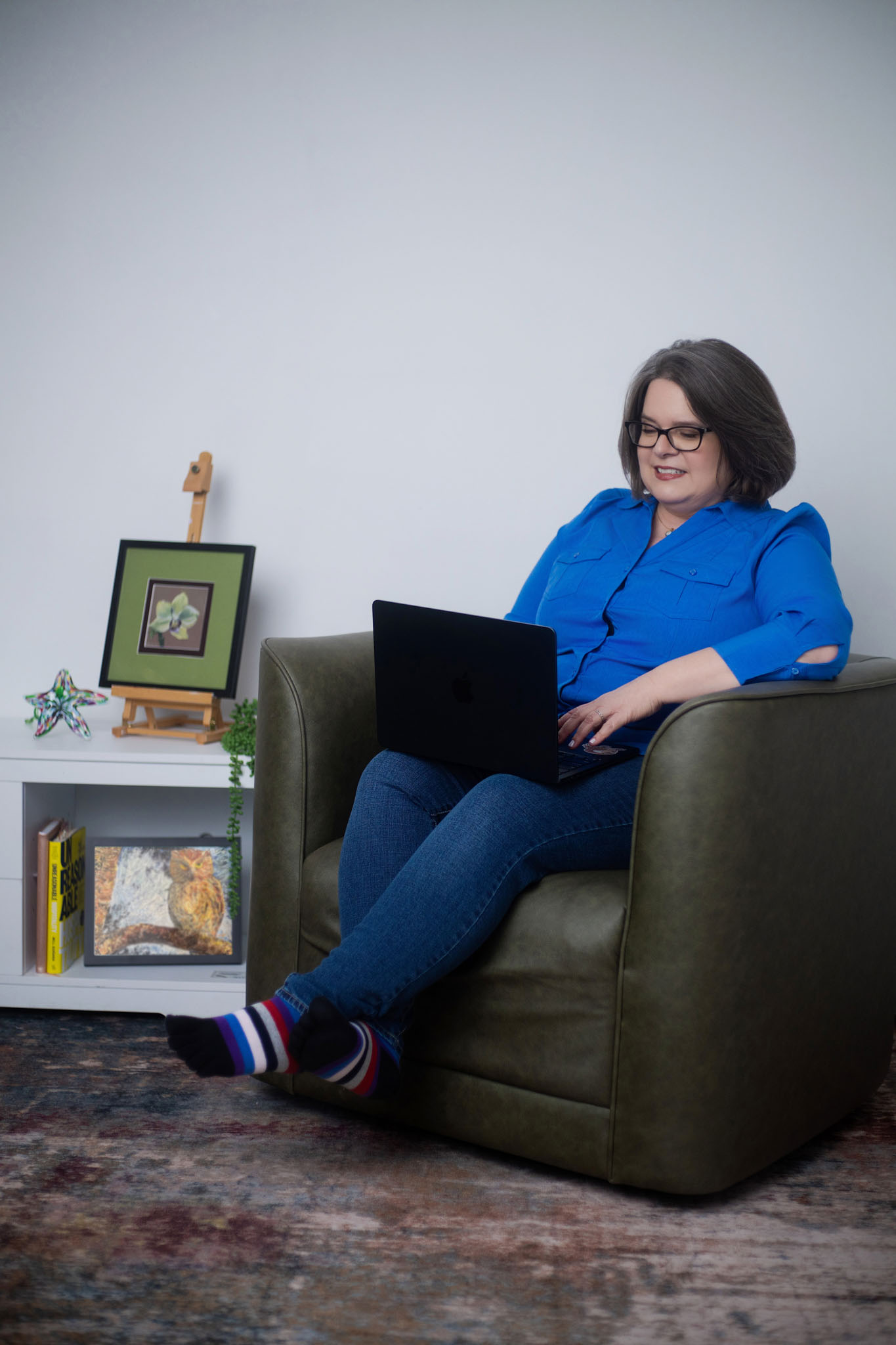 Laurie McAleenan working on her laptop in an armchair with her framed art displayed in the background