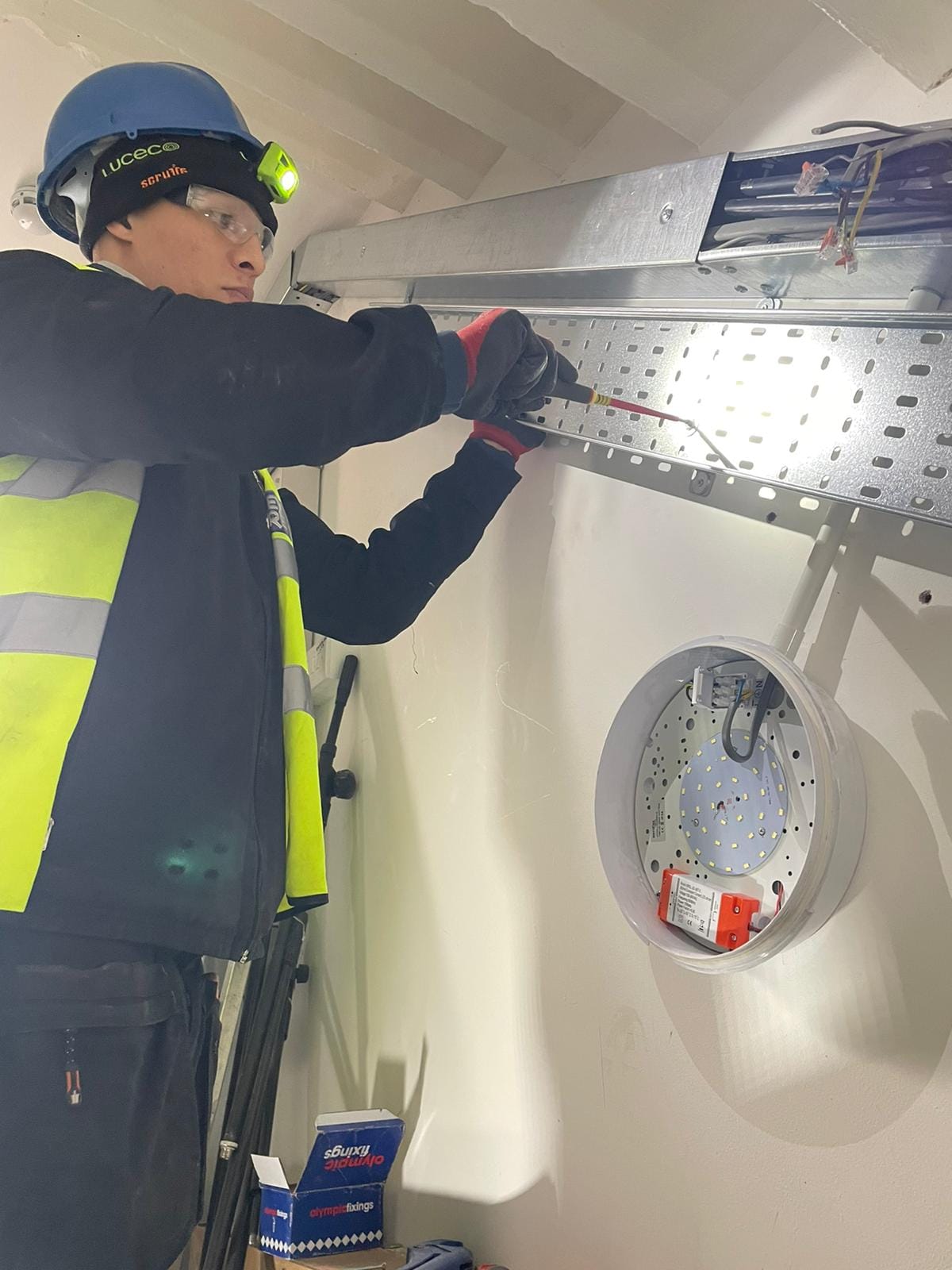Electrician in certified PPE installing and wiring an LED emergency luminaire bulkhead fitting with battery backup