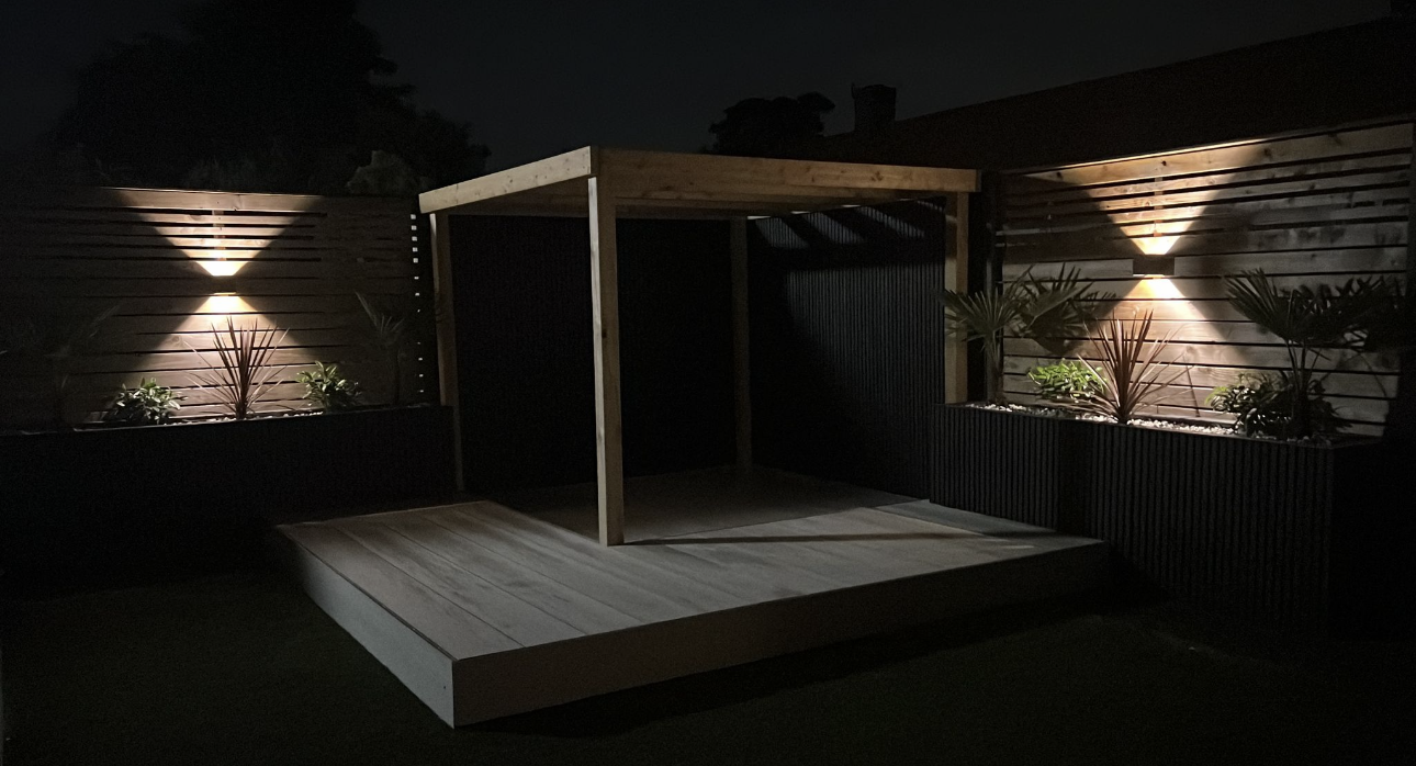 Garden room pergola deck structure with warm exterior fence sconces and planter uplighting at night