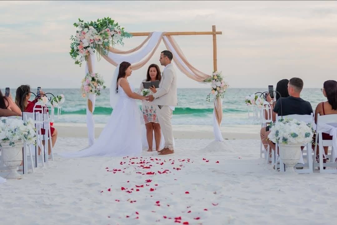 Marlene officiating an intimate vow exchange under a draped bamboo arch with rose petals on white sand