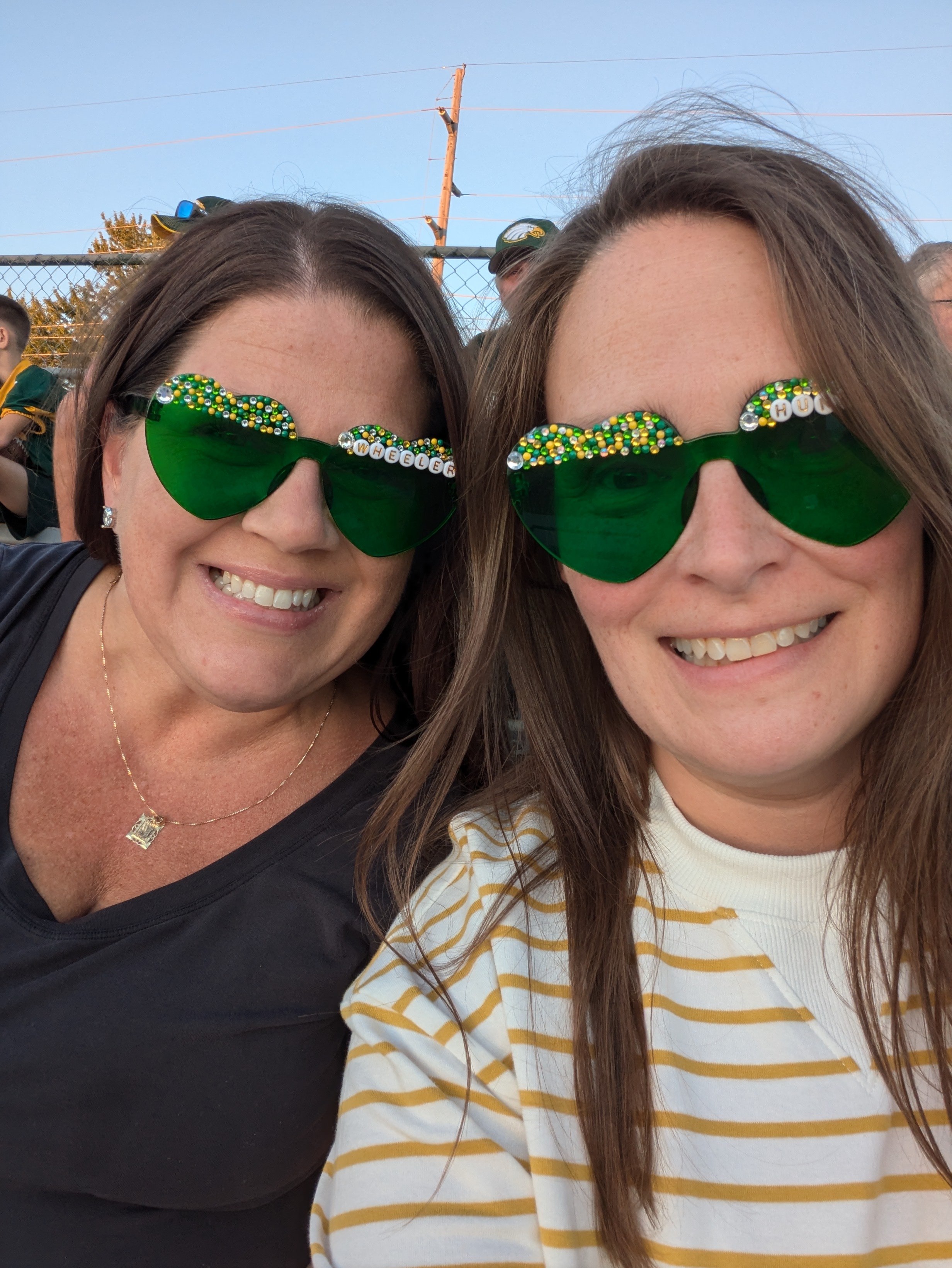 Two women smiling at a game wearing green diamond-painted sunglasses with letter personalization