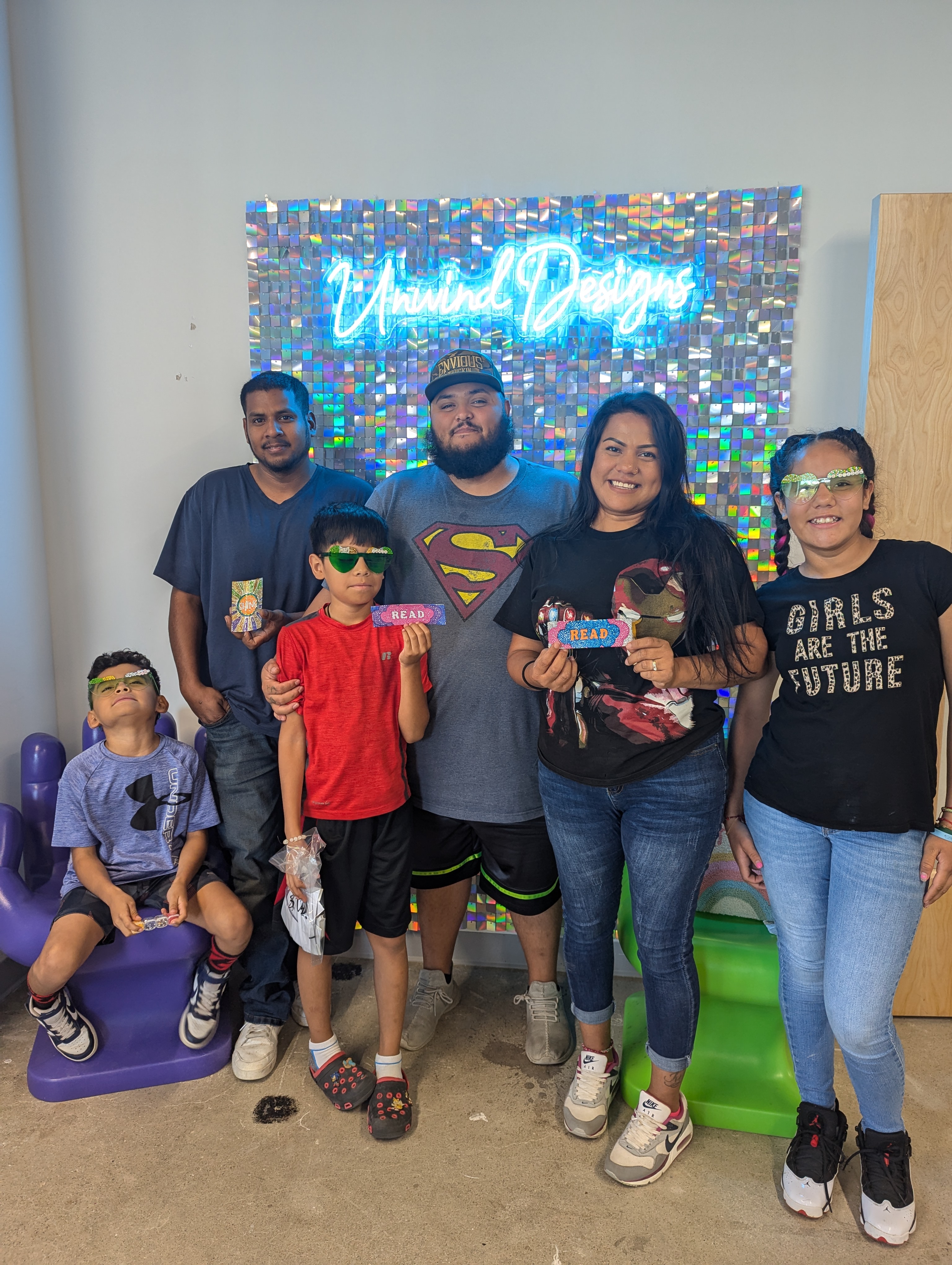 Family posing in front of the sparkling neon Unwind Designs sign with their diamond-painted creations