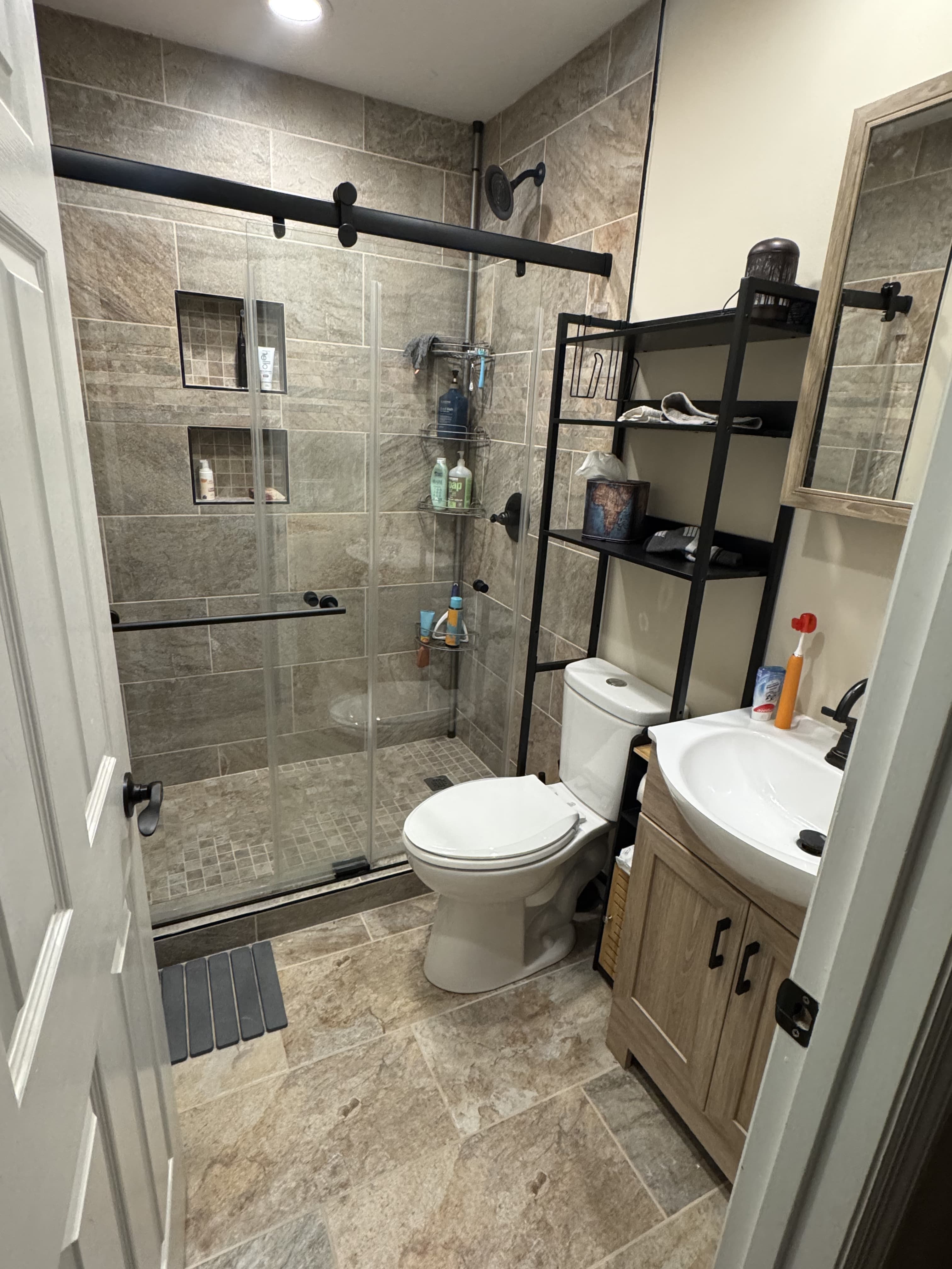 Custom walk-in shower with large-format earth tone stone tile, dual recessed mosaic niches, and matte black sliding glass barn door by Southern Forge Construction