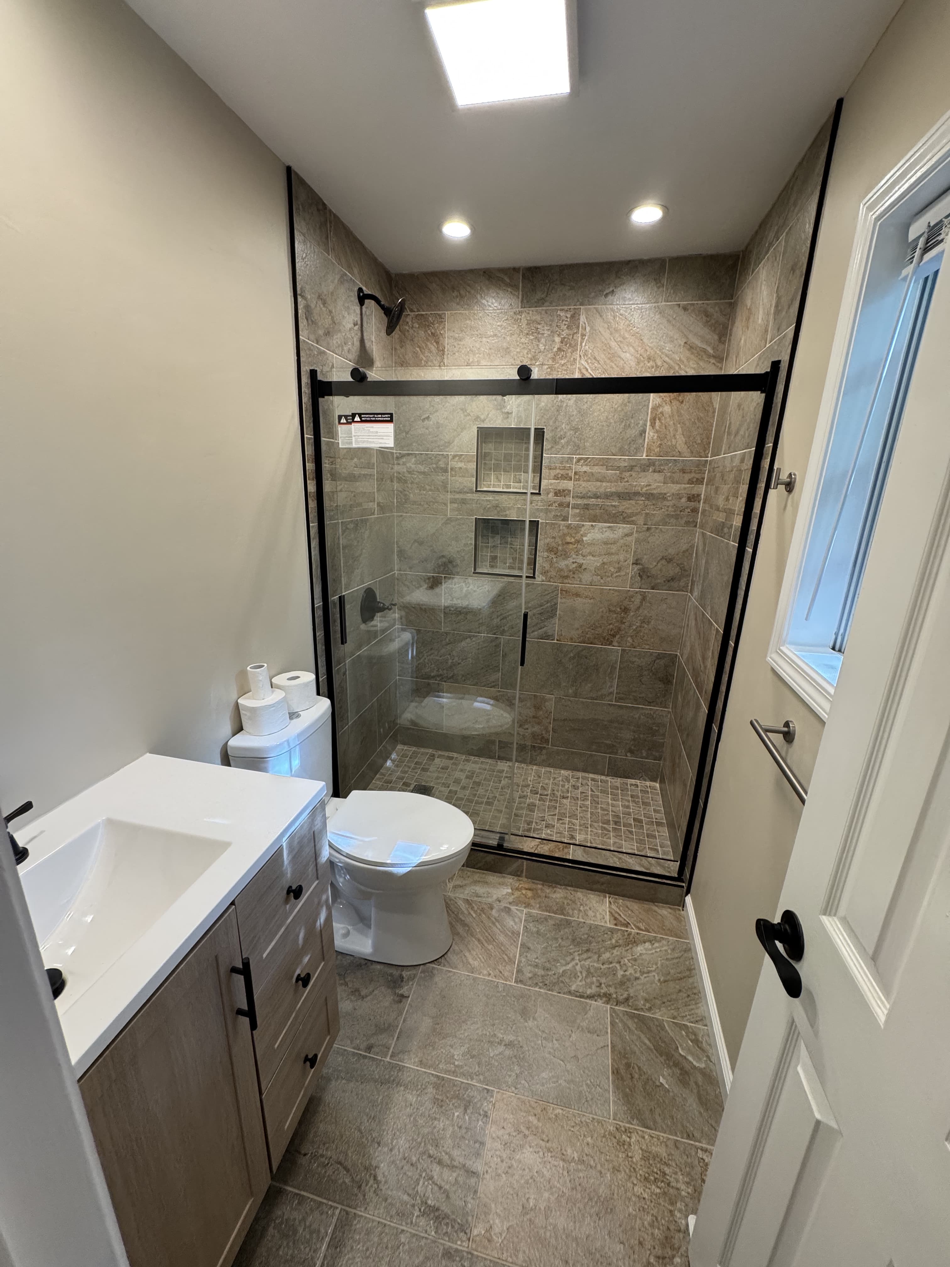 Finished bathroom renovation with custom light wood vanity, white quartz sink top, black hardware, and fully tiled glass shower enclosure by Southern Forge Construction