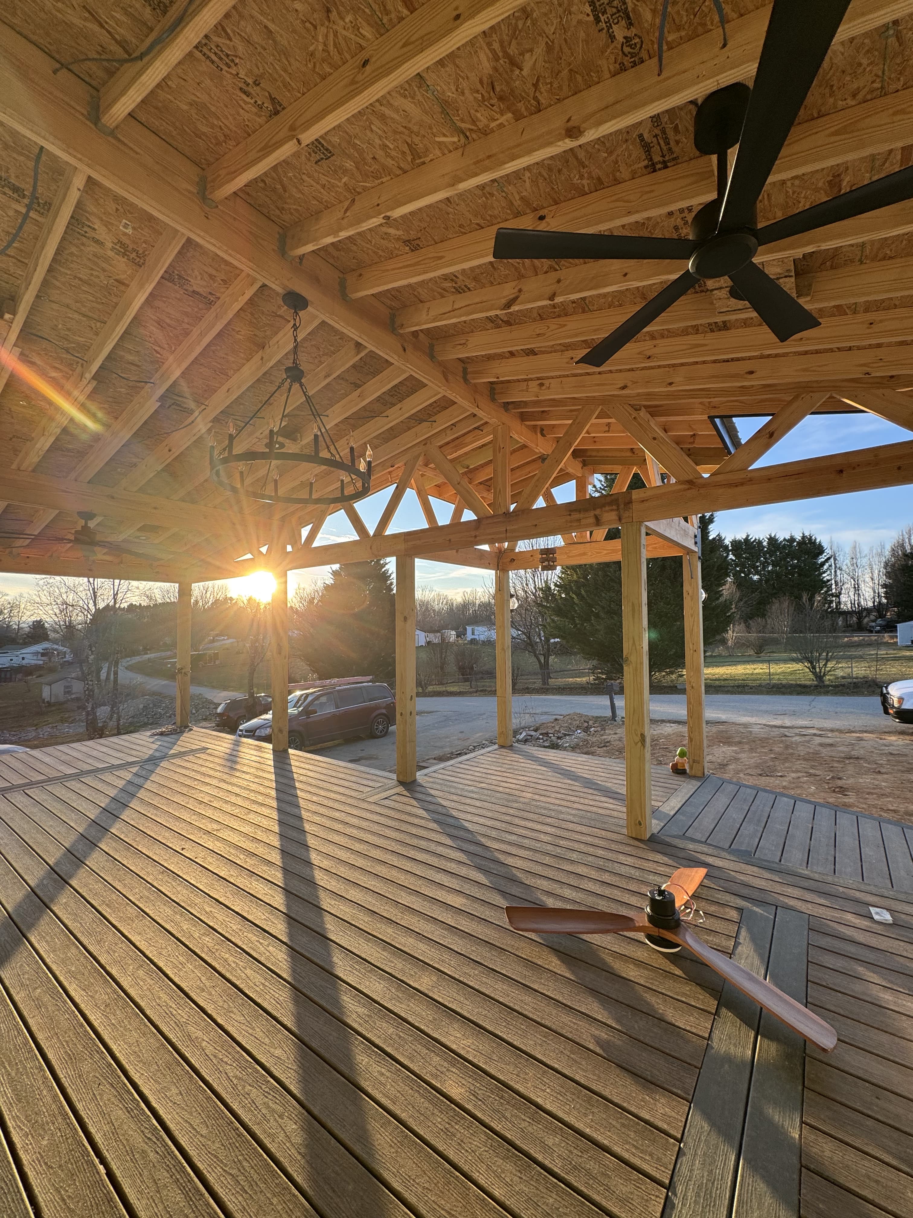 Underside of vaulted timber truss roof with exposed wood rafters, wagon wheel chandelier, and wide composite deck floor at sunset