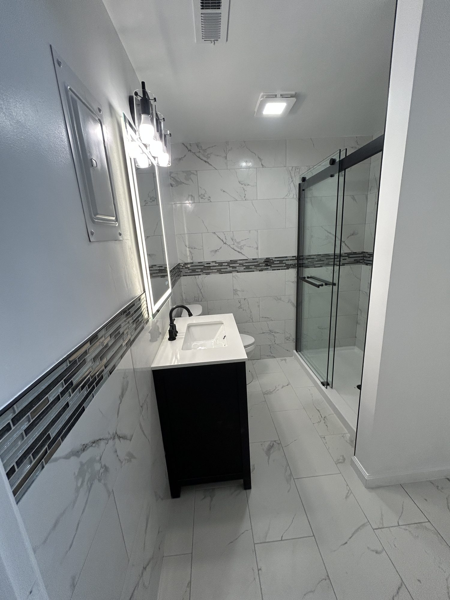 Long view of bathroom remodel showing navy vanity, lit mirror, marbled tile, and glass shower, Reno, NV