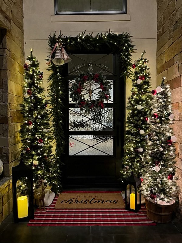 Illuminated evening Christmas porch installation in San Antonio with lighted evergreen trees, custom door garland, bells, and holiday lanterns