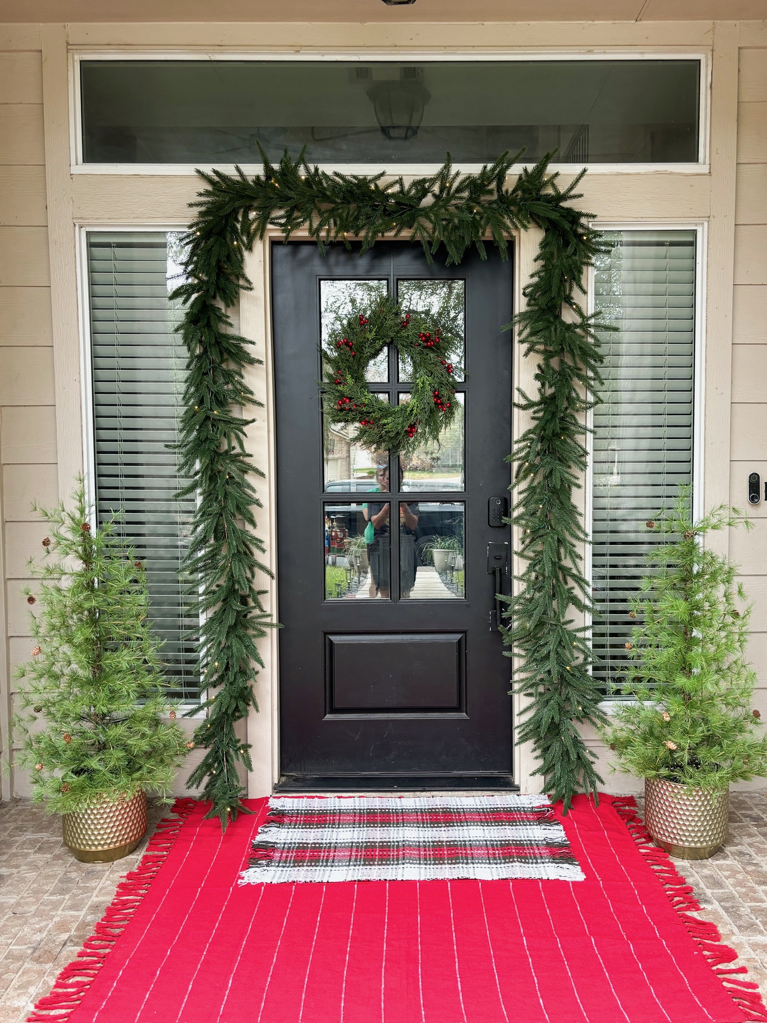 Holiday front porch with lush cedar door garland, red plaid welcome rug, door wreath with berries, and flanking potted pine trees in San Antonio