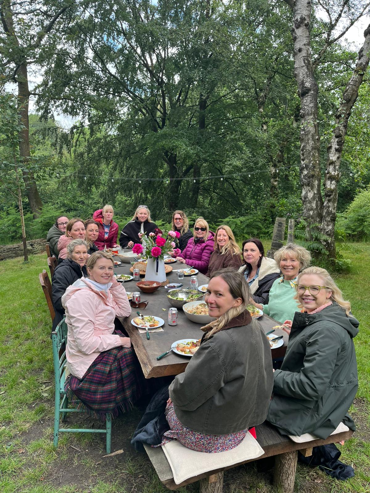 Women gathered around a long rustic table having lunch together in the woods at the Summer Retreat