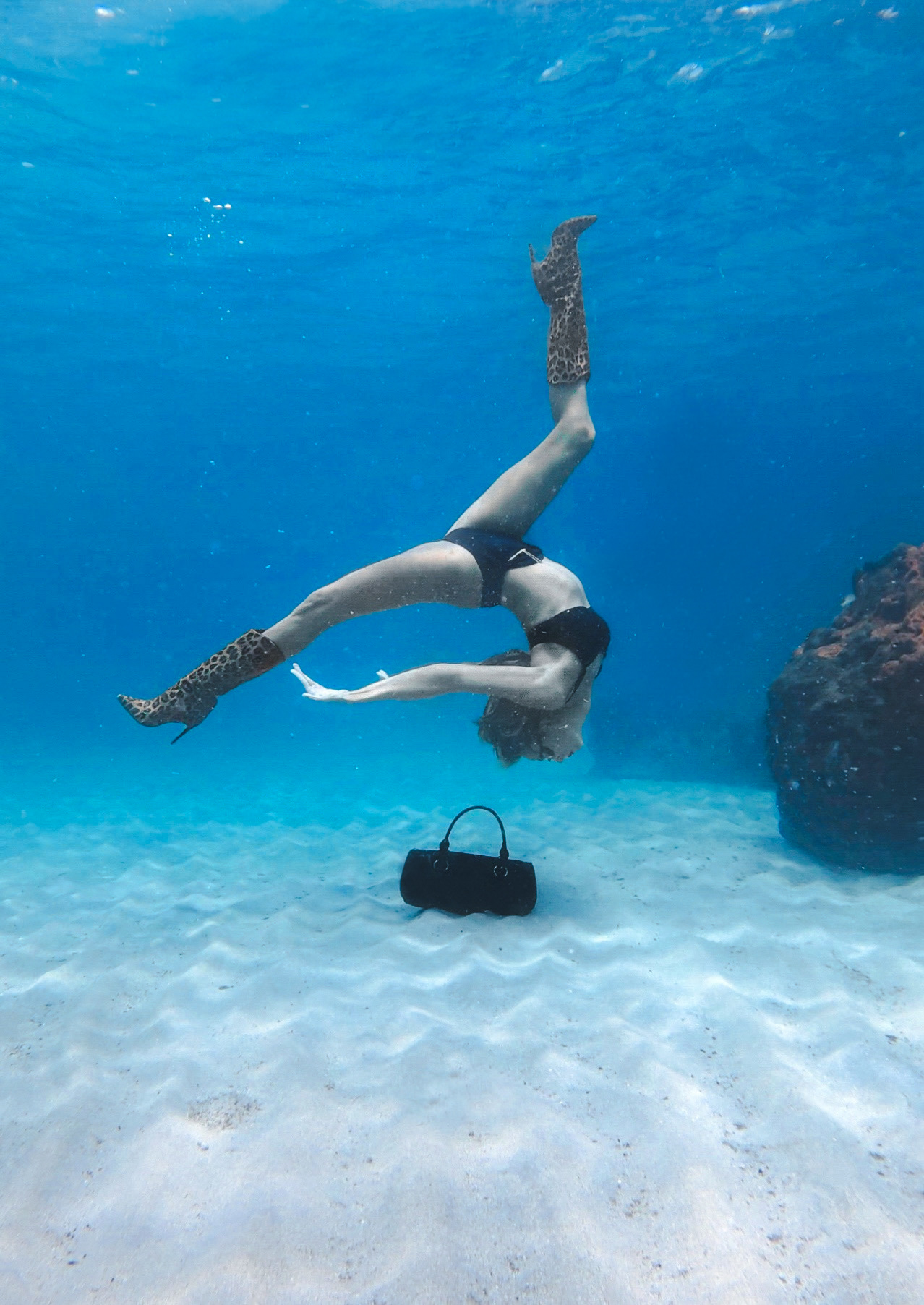 Kristina Makushenko upside down underwater beside a handbag on the sand