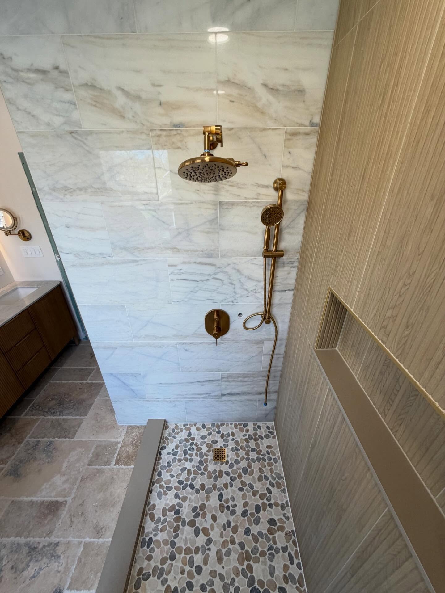 Close-up of walk-in shower featuring brushed-gold rain shower head, handheld sprayer, pebble mosaic tile floor, and recessed storage niche.