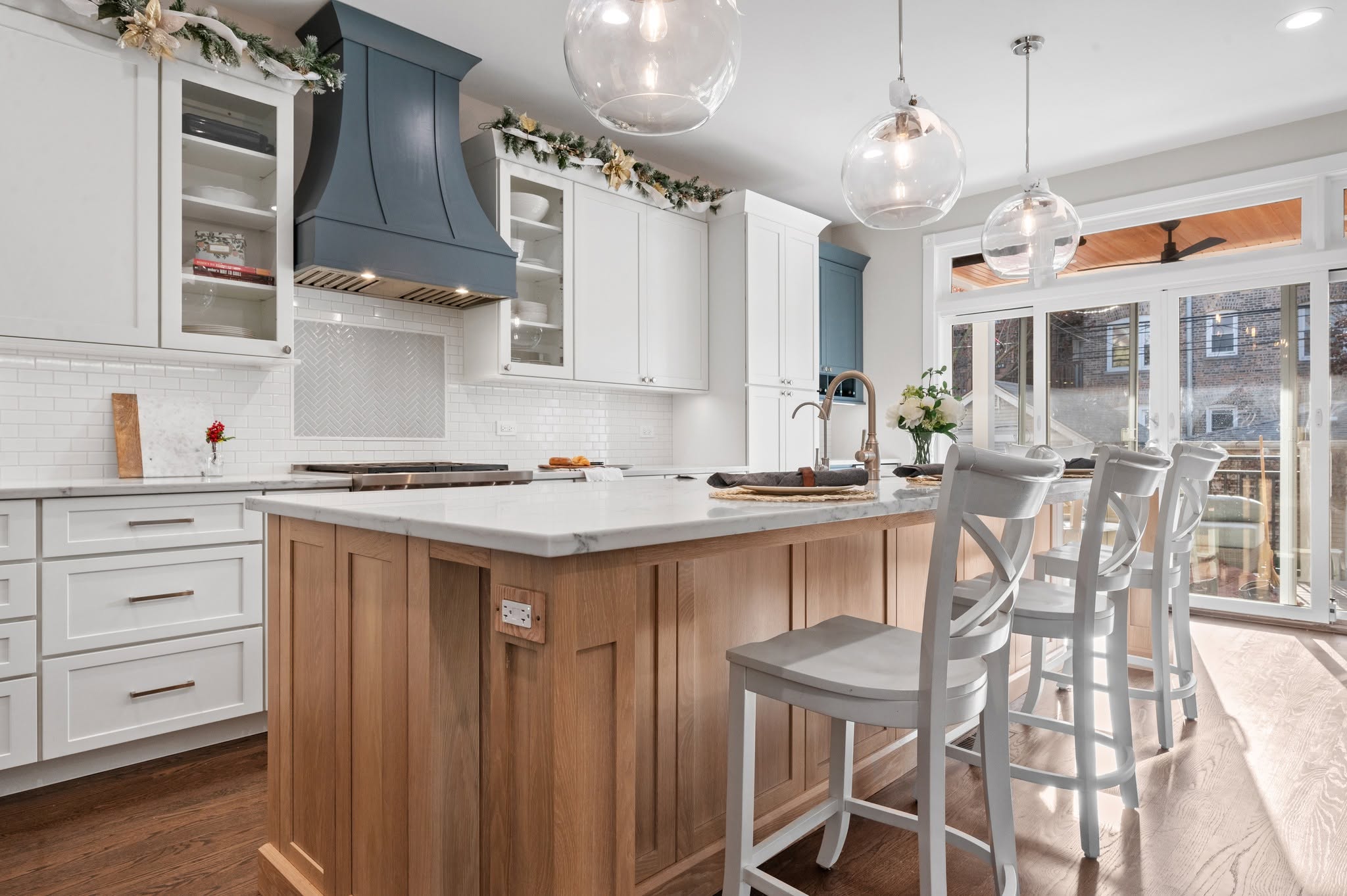View across the quartz island toward the range, custom hood, white perimeter cabinets, and glass upper storage.