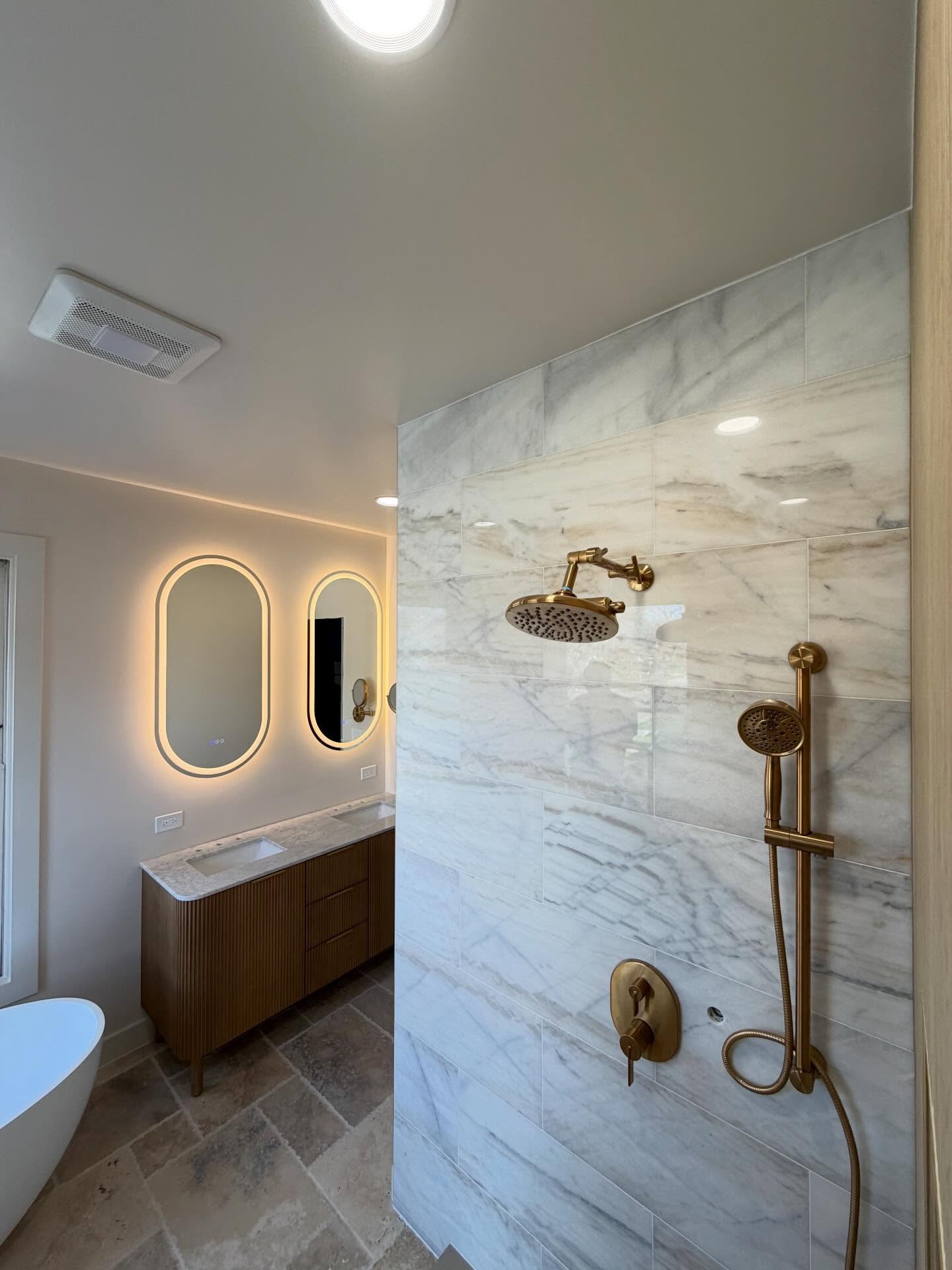 Perspective across marble shower wall toward double floating vanity with backlit illuminated mirrors and warm lighting.