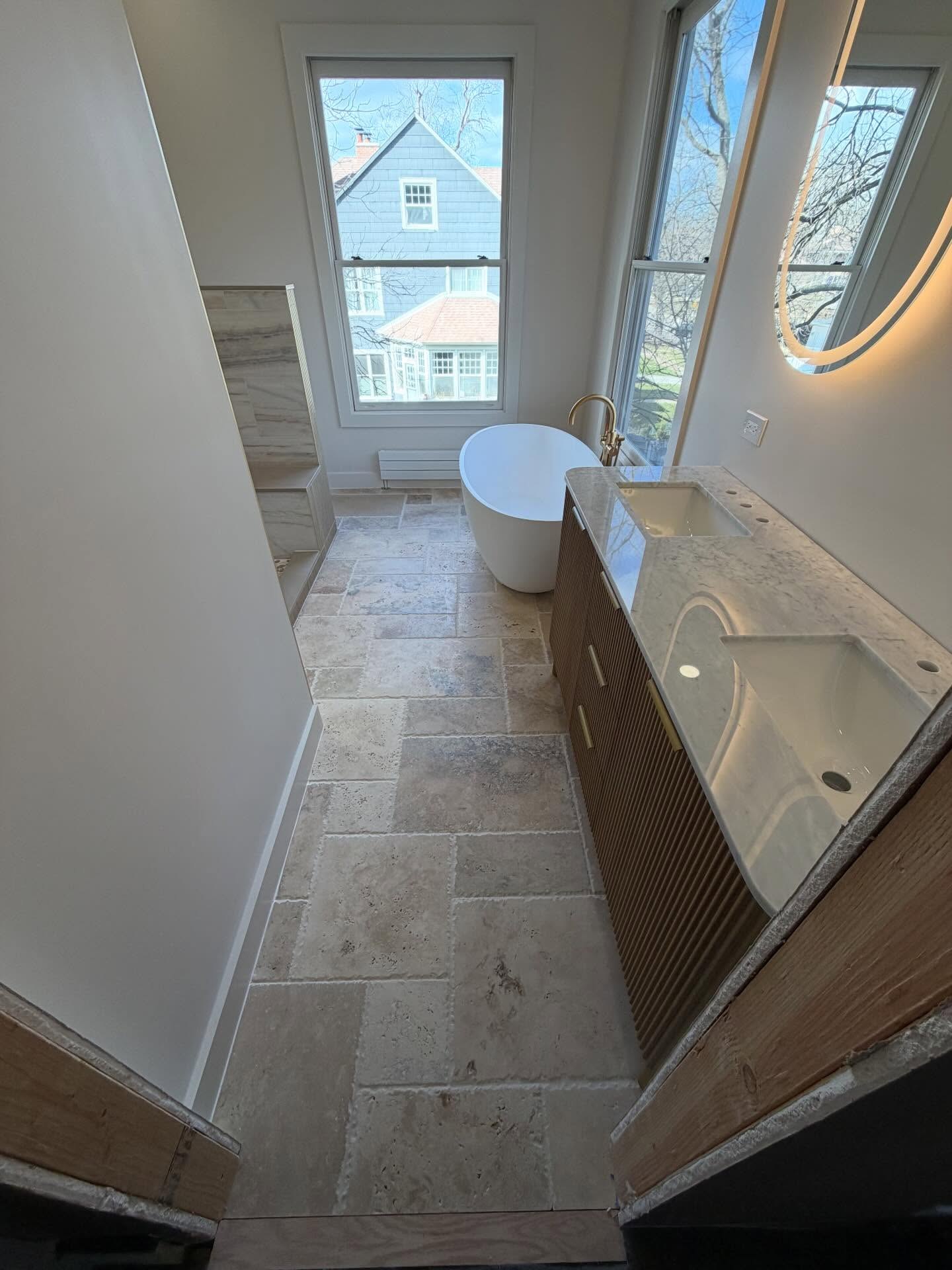 Full view of warm contemporary primary bathroom with natural stone-look floor tile, freestanding tub, and fluted double vanity.