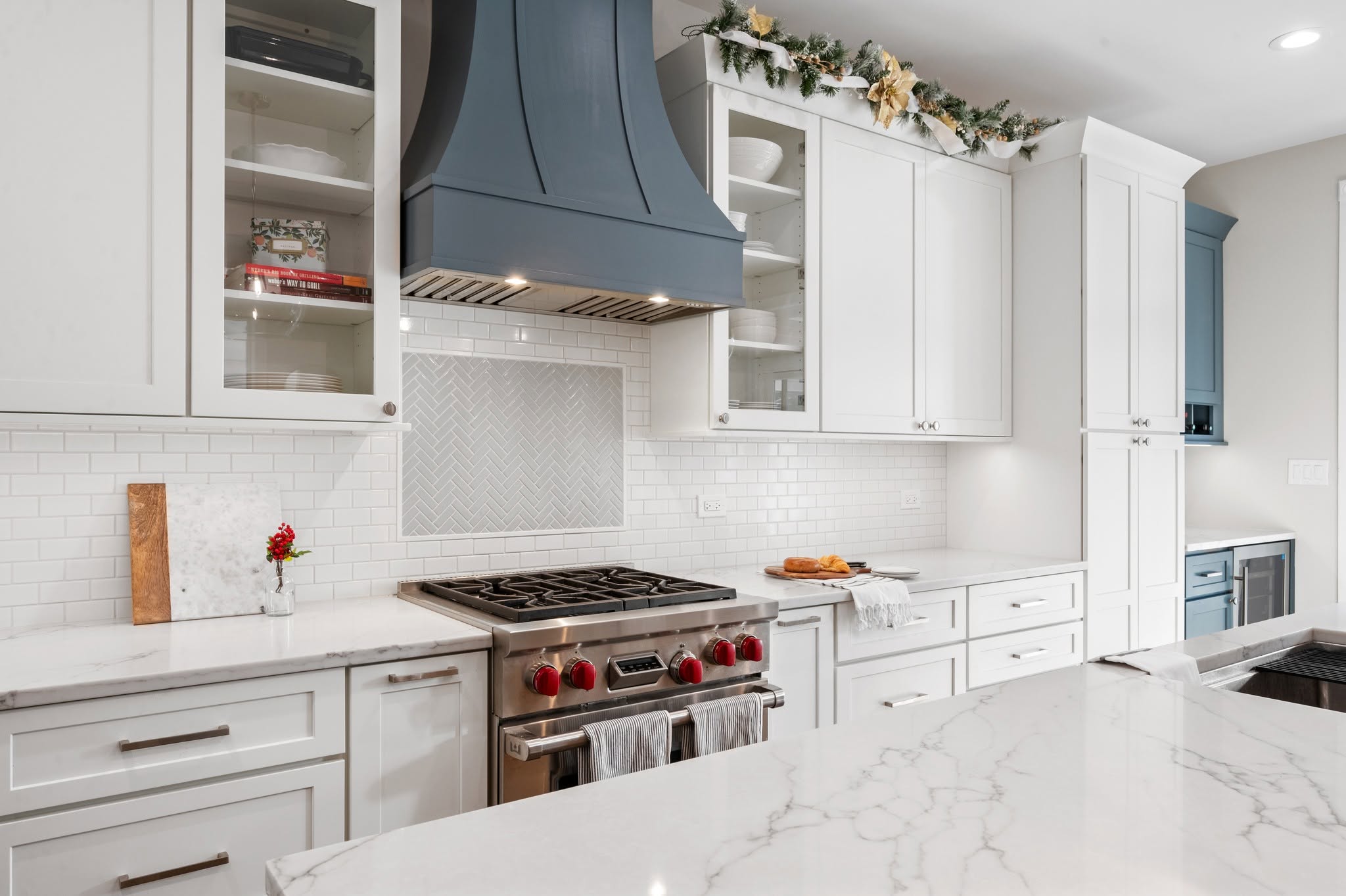 Custom dark range hood with integrated lighting, herringbone and subway tile backsplash, commercial-style range, and glass-front upper cabinets.