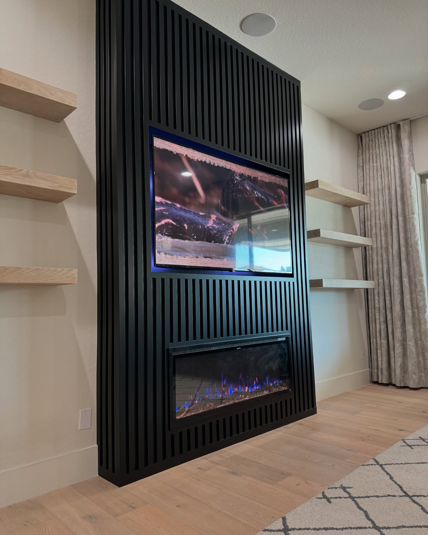 Angled perspective showing the black vertical slat texture, modern electric fireplace, and warm natural wood floating shelves.