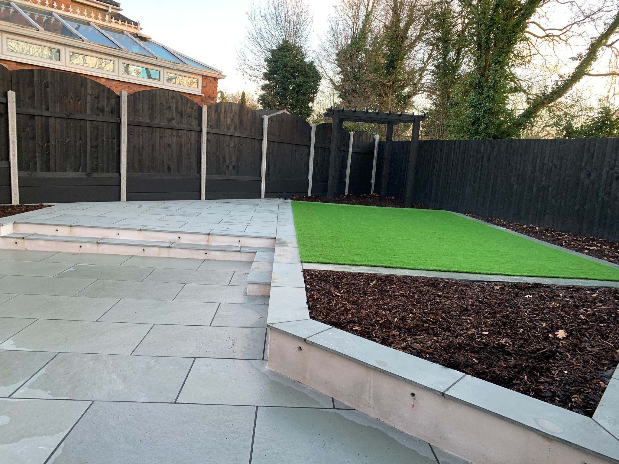Neat artificial grass lawn with raised patio and bark chip garden border in Essex