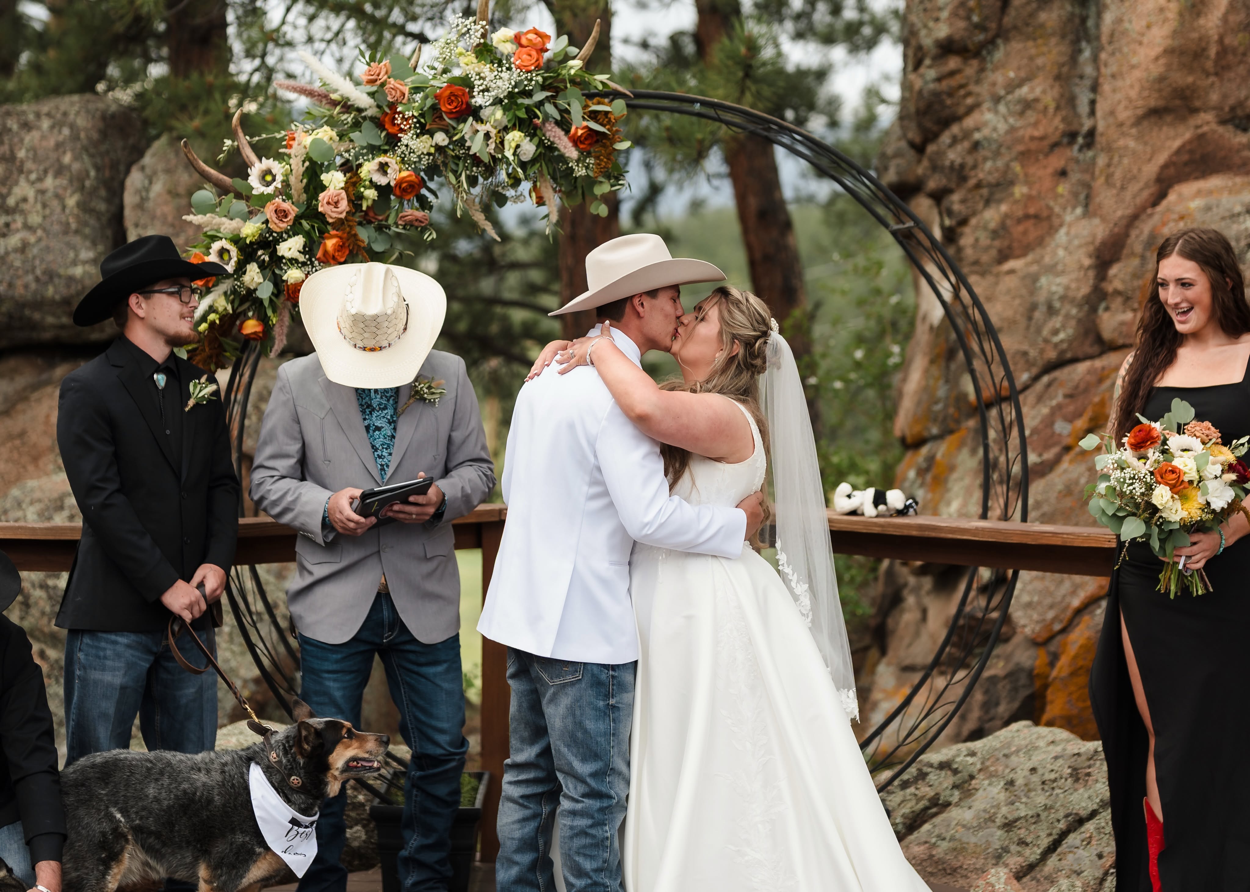 A bride and groom share their first kiss beneath a floral arch framed by Colorado pines and rock formations at Deer Creek Valley Ranch