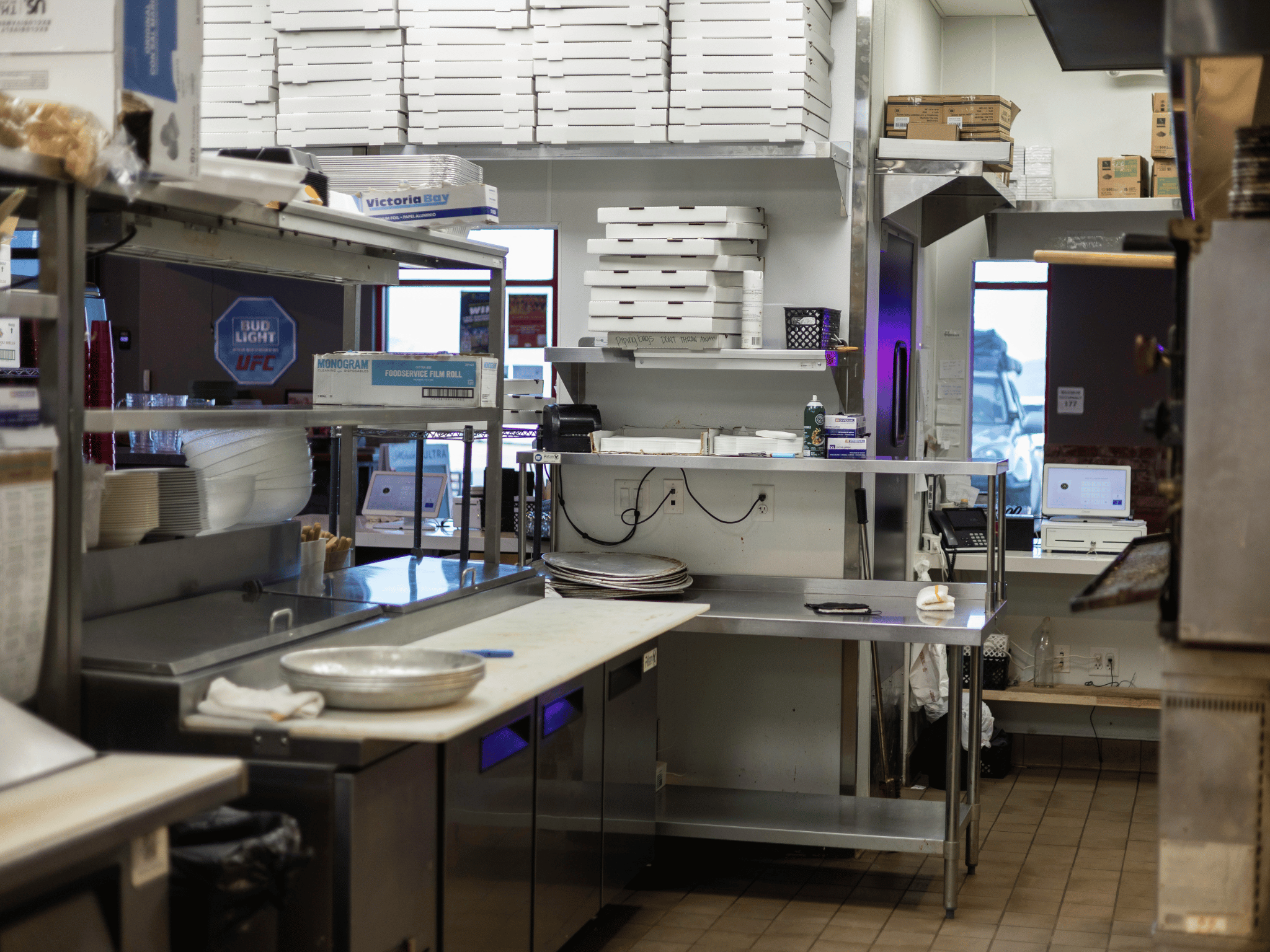 Commercial kitchen prep line with stainless steel worktables, refrigeration, and pizza box storage racks