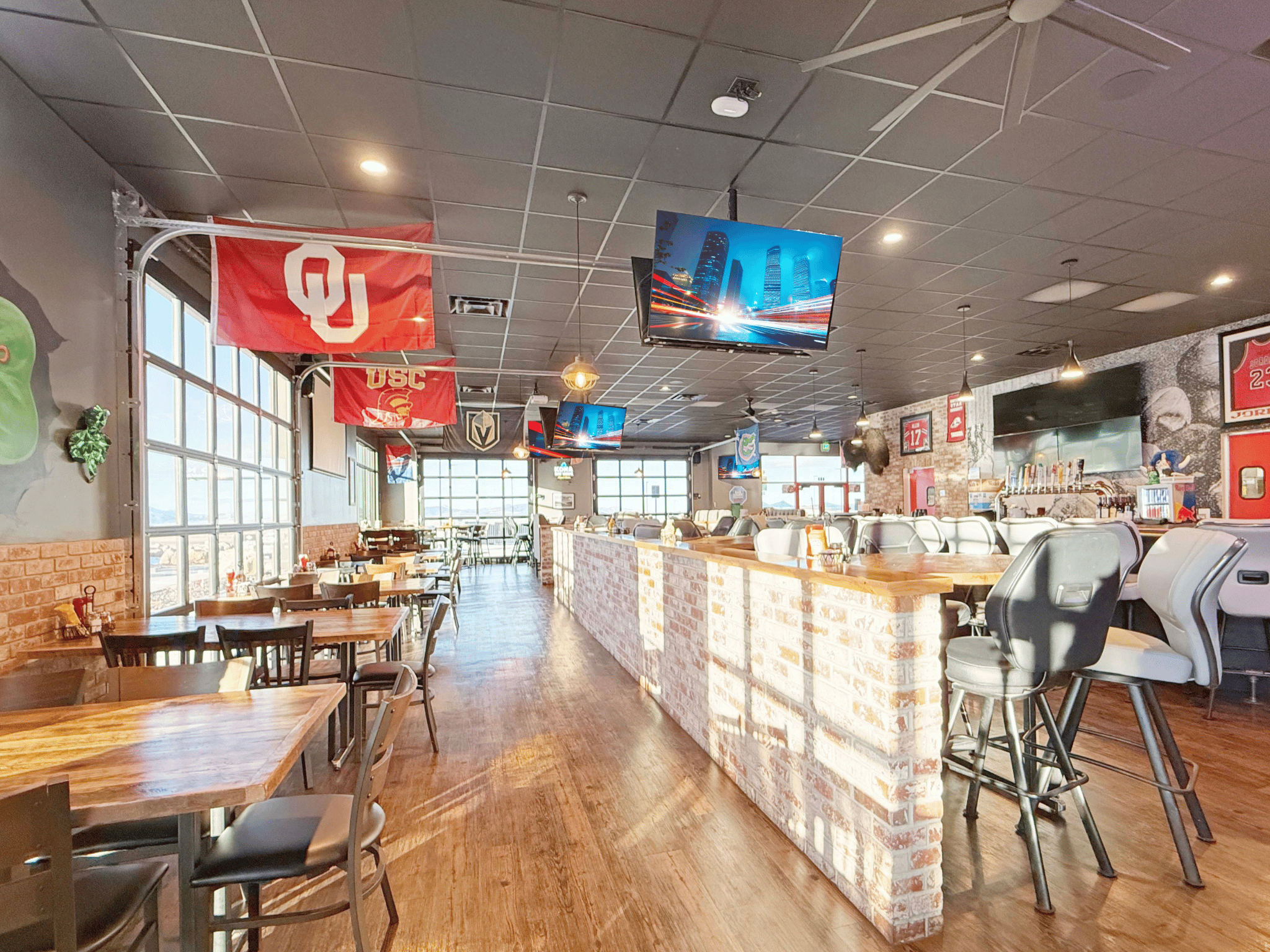 Spacious dining room and bar area with brick half-wall, wooden tables, overhead monitors, and glass garage doors