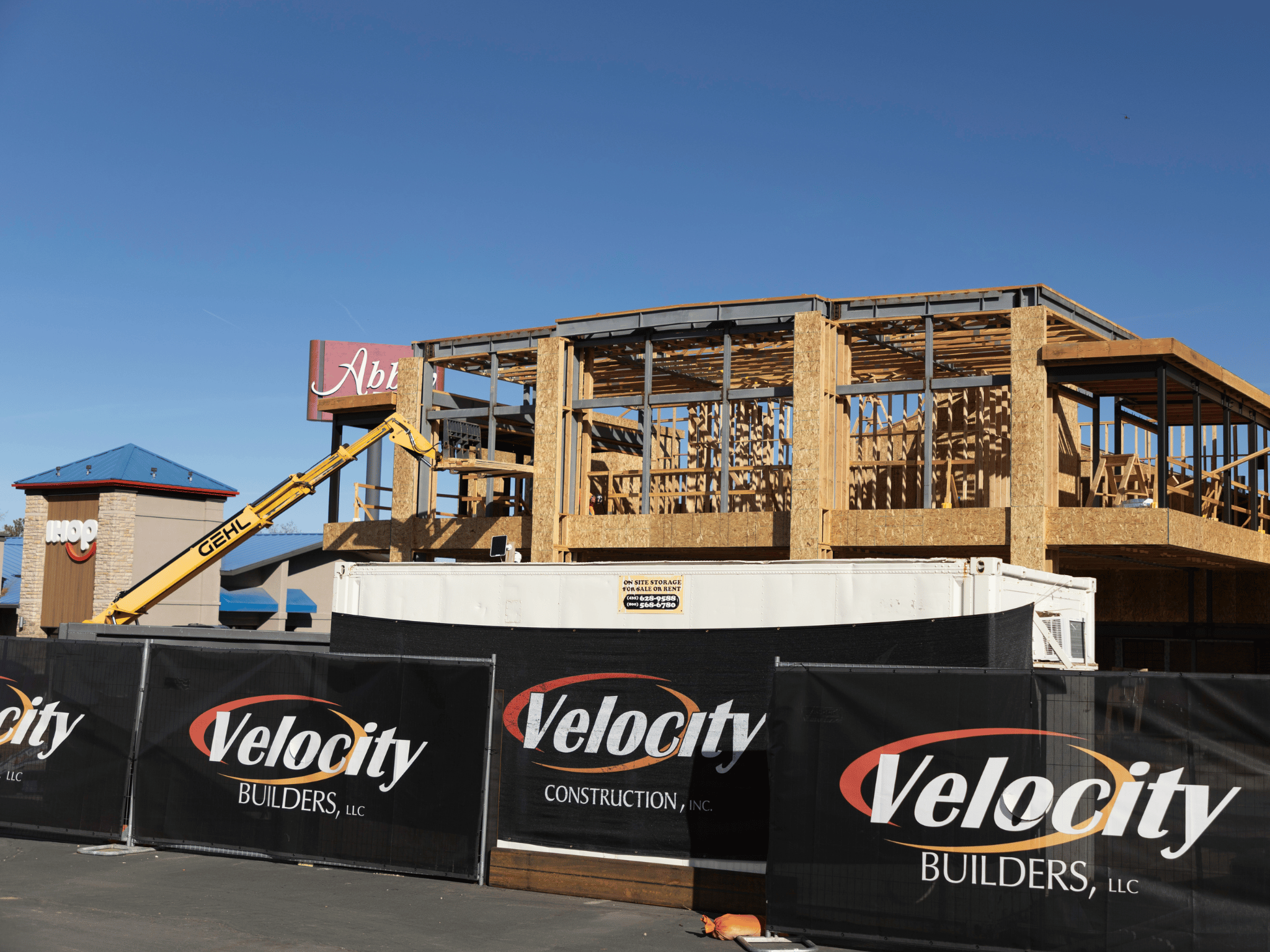 Active commercial construction site with structural steel and wood framing, telehandler equipment, and Velocity construction fencing