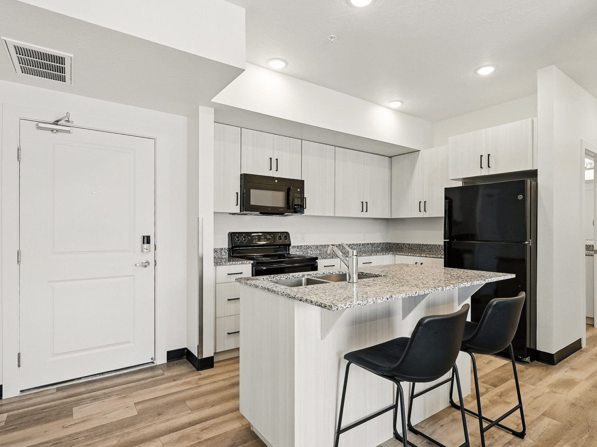 Modern student apartment kitchen with white cabinetry, granite island counter, barstools, and black appliances
