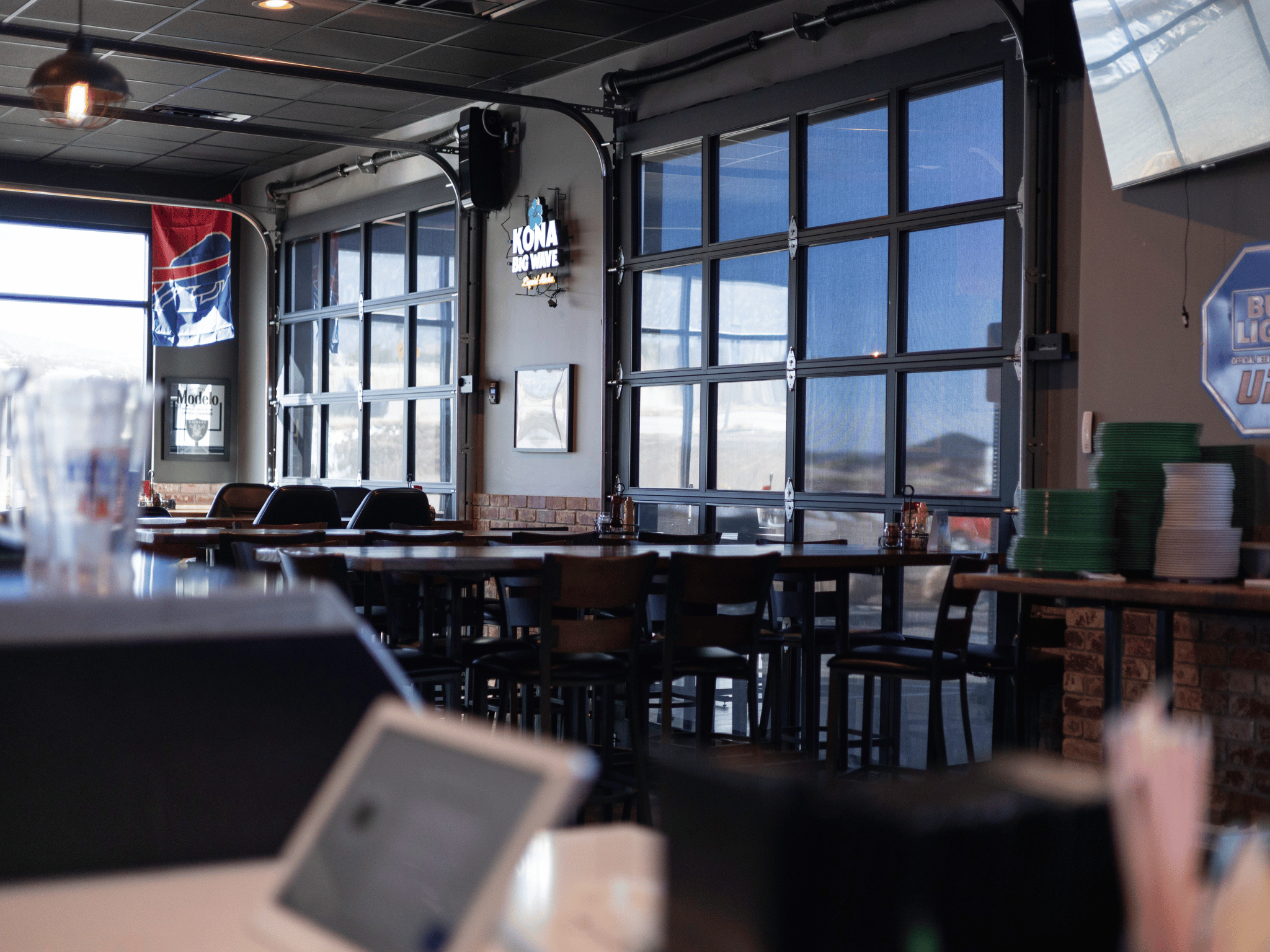 Dining tables positioned along glass roll-up patio garage doors with neon signs and natural lighting