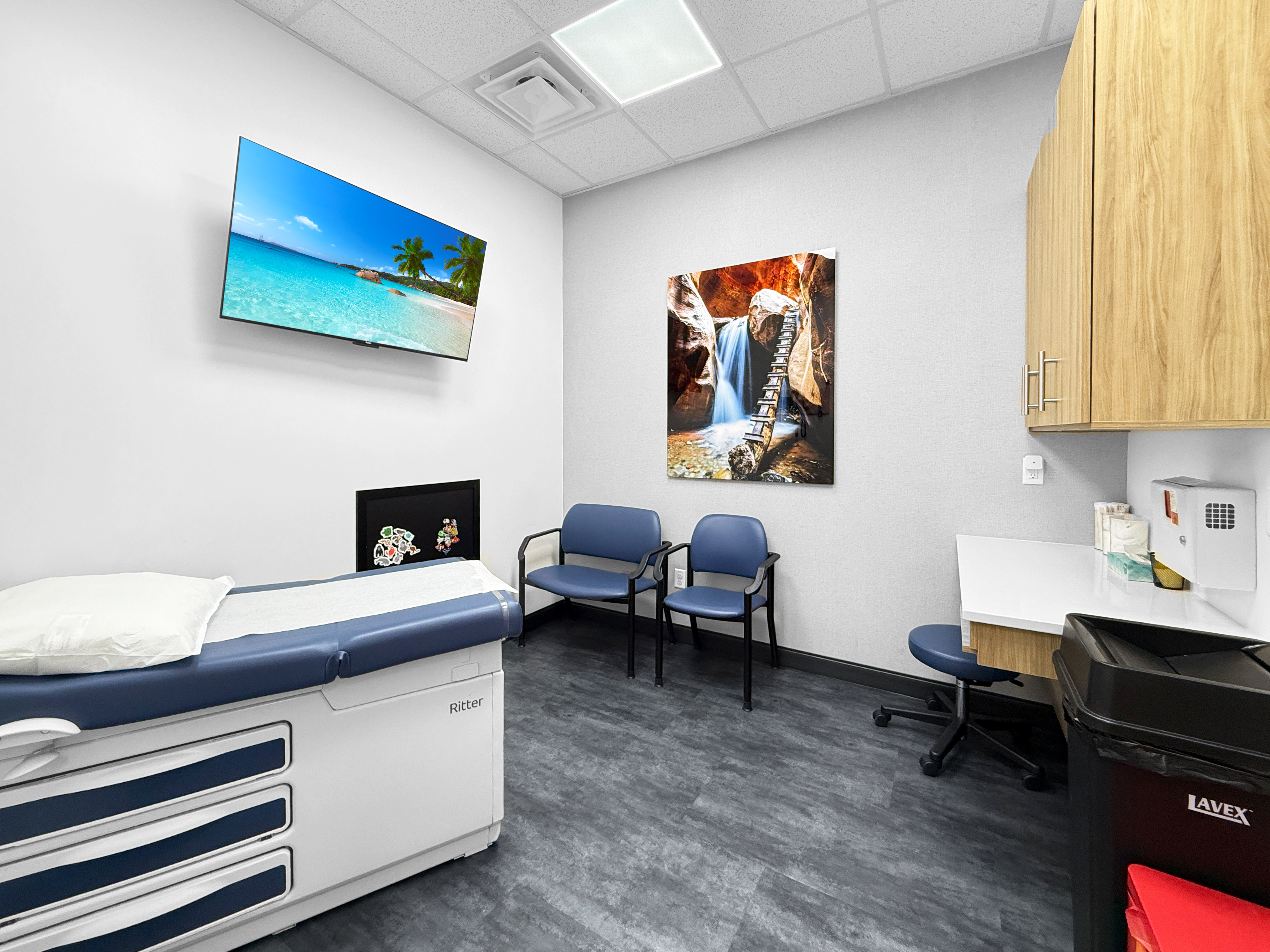 Patient examination room with exam table, patient seating, digital display, and medical cabinetry