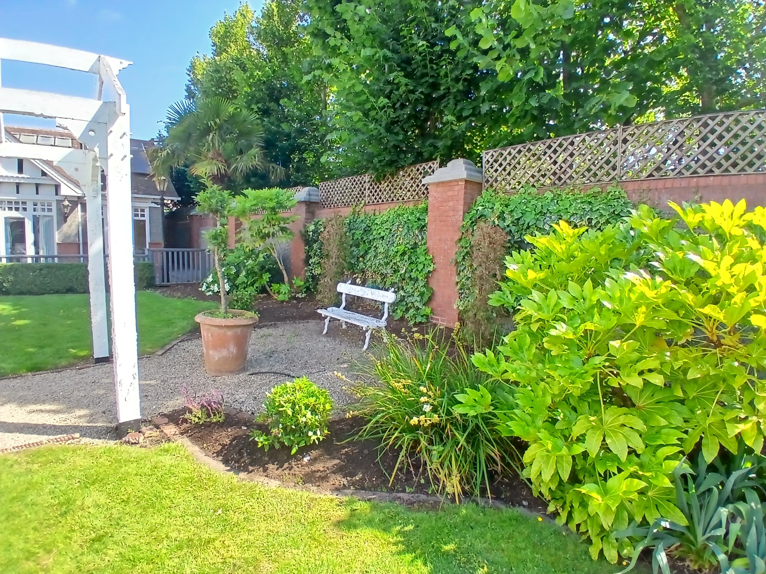 Formal garden courtyard featuring white timber pergola, terracotta planter, garden bench, brick piers with trellis fencing and lush surrounding planting.