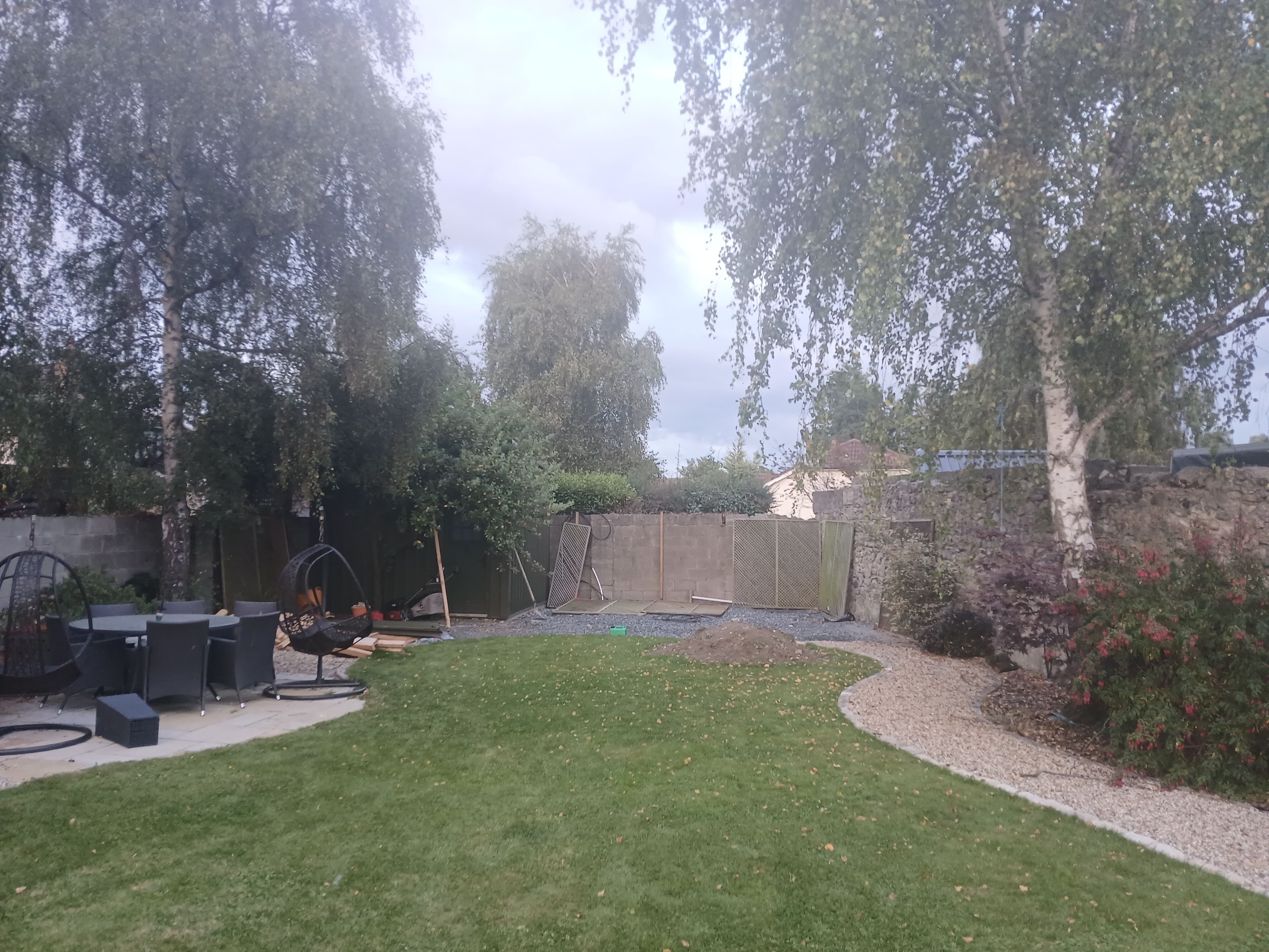 Wide panoramic garden overview showing lawn, curved gravel path, seating patio and mature birch trees.