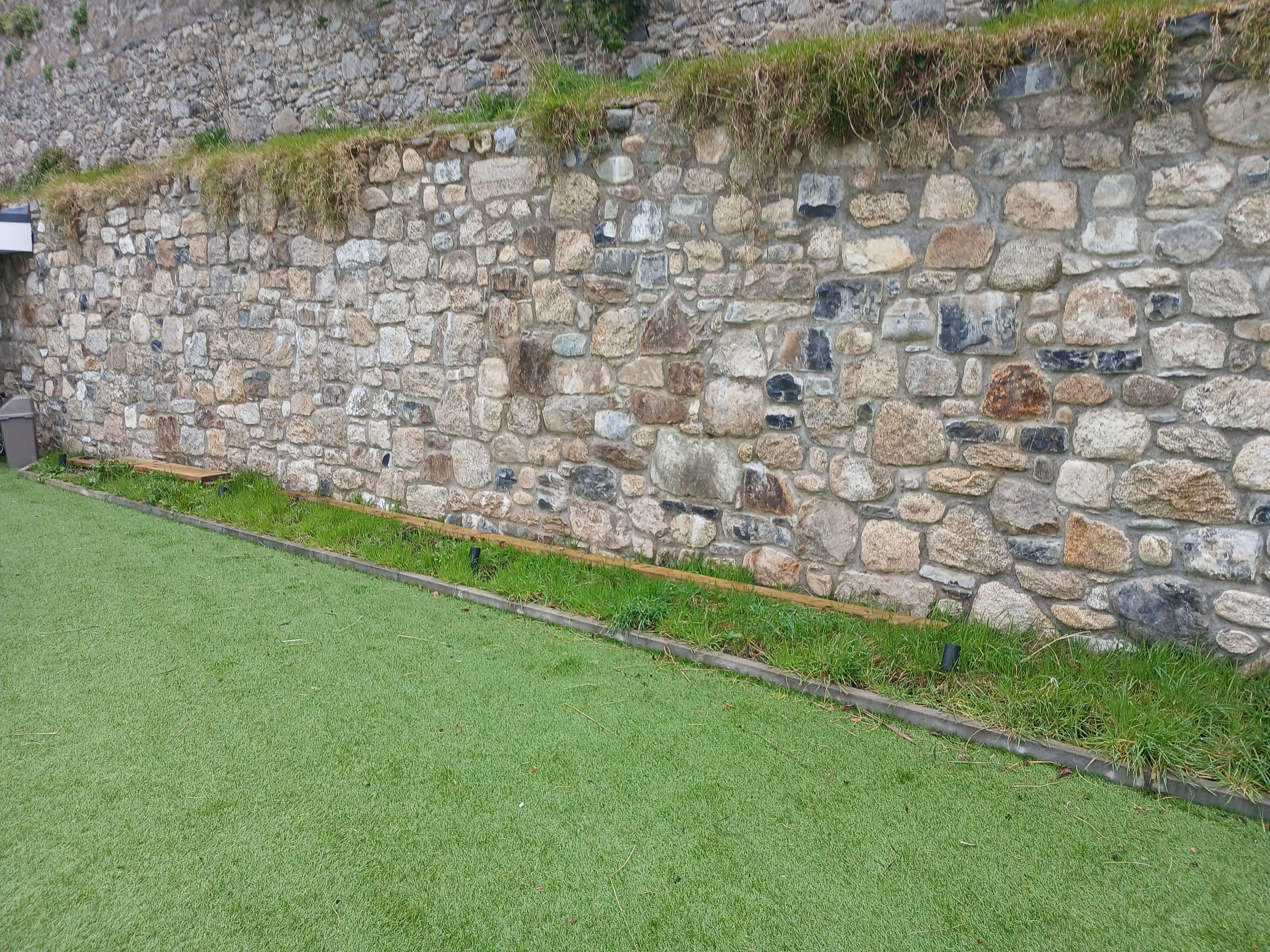 Lawn boundary edged neatly with treated timber edging and fresh turf alongside a historic granite stone boundary wall.