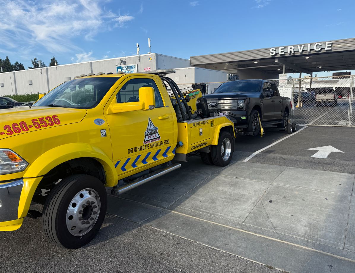 Pure Towing & Recovery bright yellow Ram 5500 tow truck with 1051 Richard Ave Santa Clara address and logo towing a black Ford F-150 Lightning into a dealership service lane