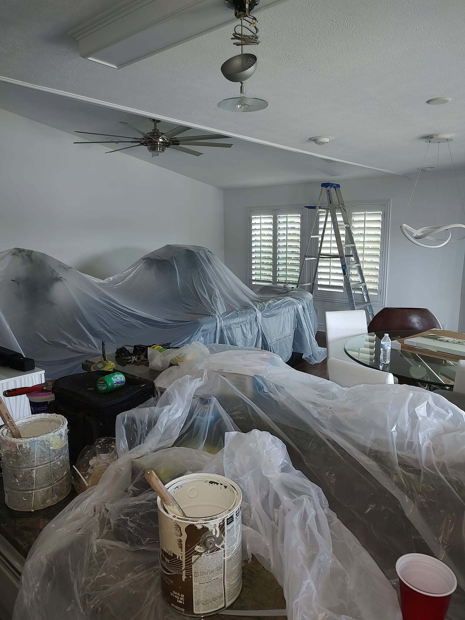 Living and dining furniture fully covered with plastic draping, paint cans staged, and stepladder ready for interior wall painting in Naples