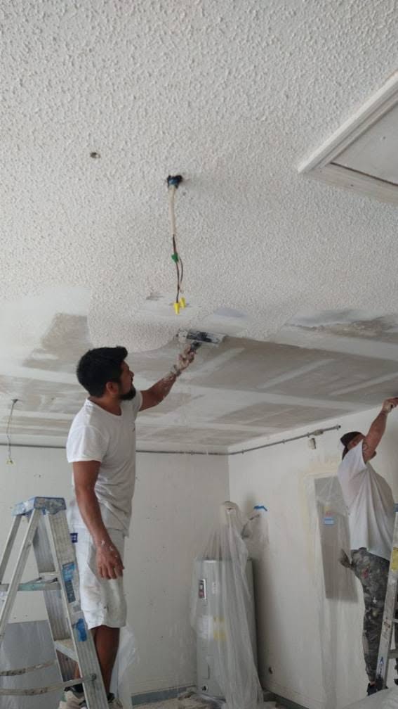Two painters scraping popcorn texture down to drywall substrate with poly masking in Naples