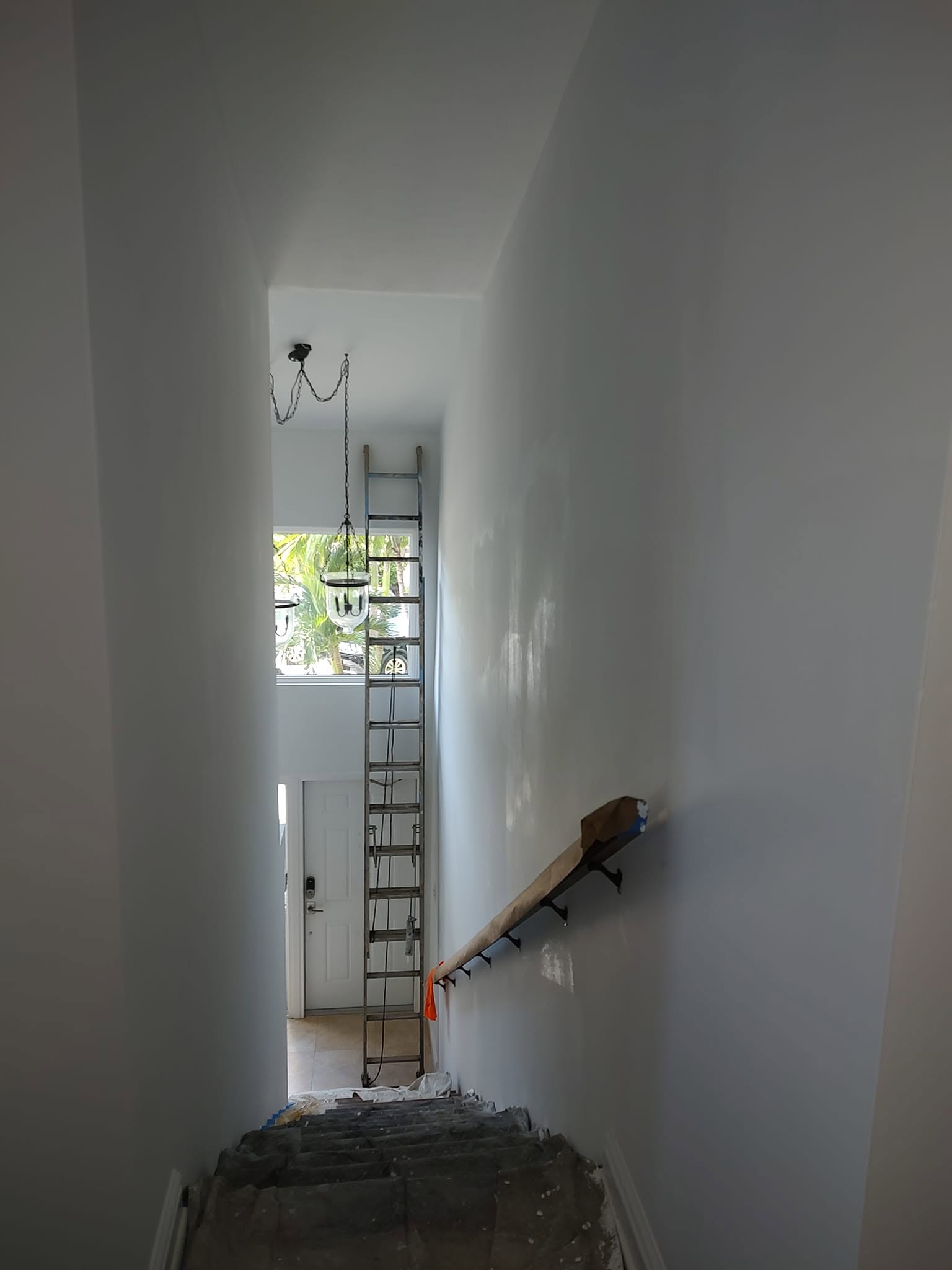 Tall extension ladder staged in narrow two-story stairwell with protected handrail and stairs for high-reach interior painting in Naples